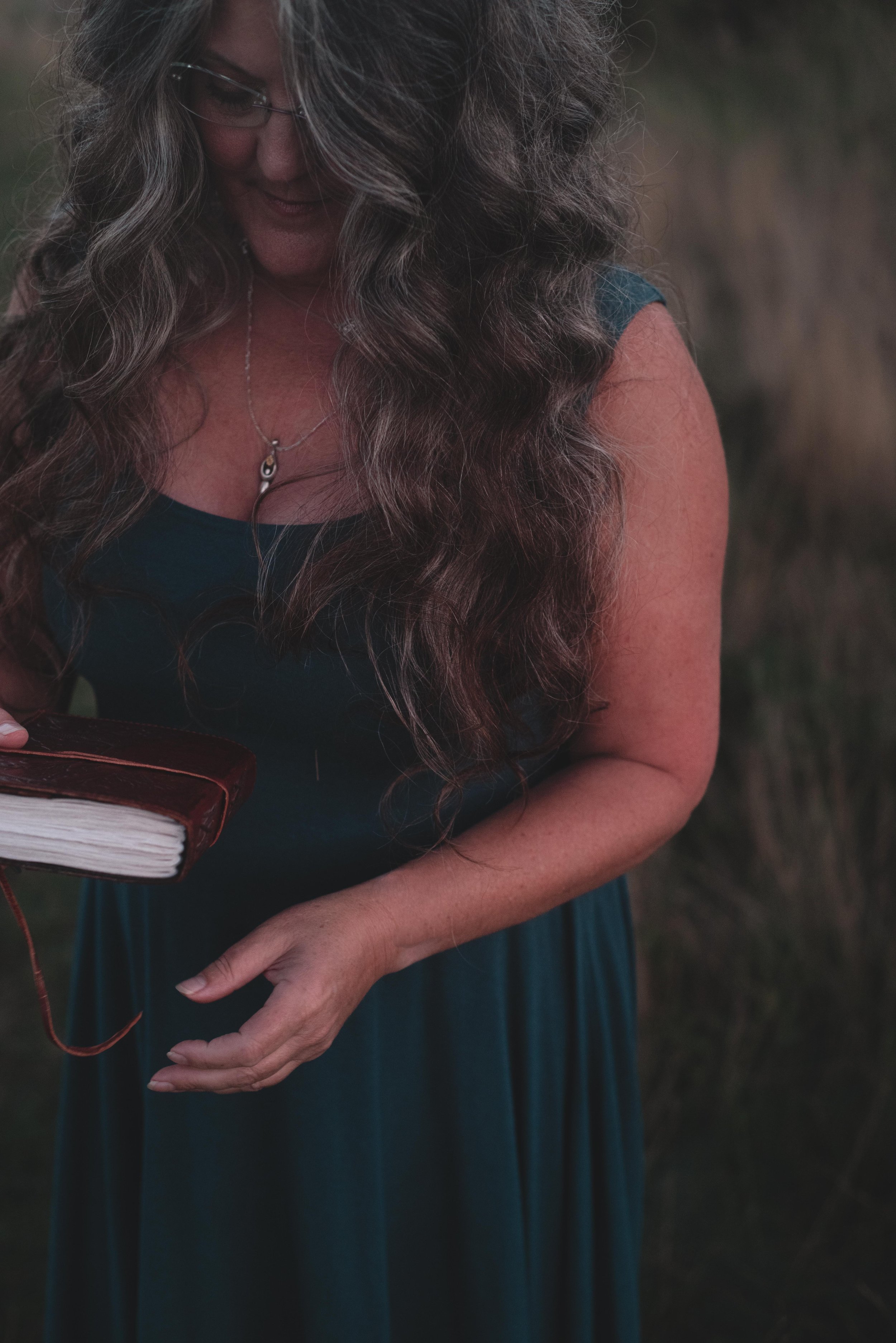 A woman with long, wavy hair holding a closed book outdoors, wearing glasses and a necklace, with a blurred natural background.