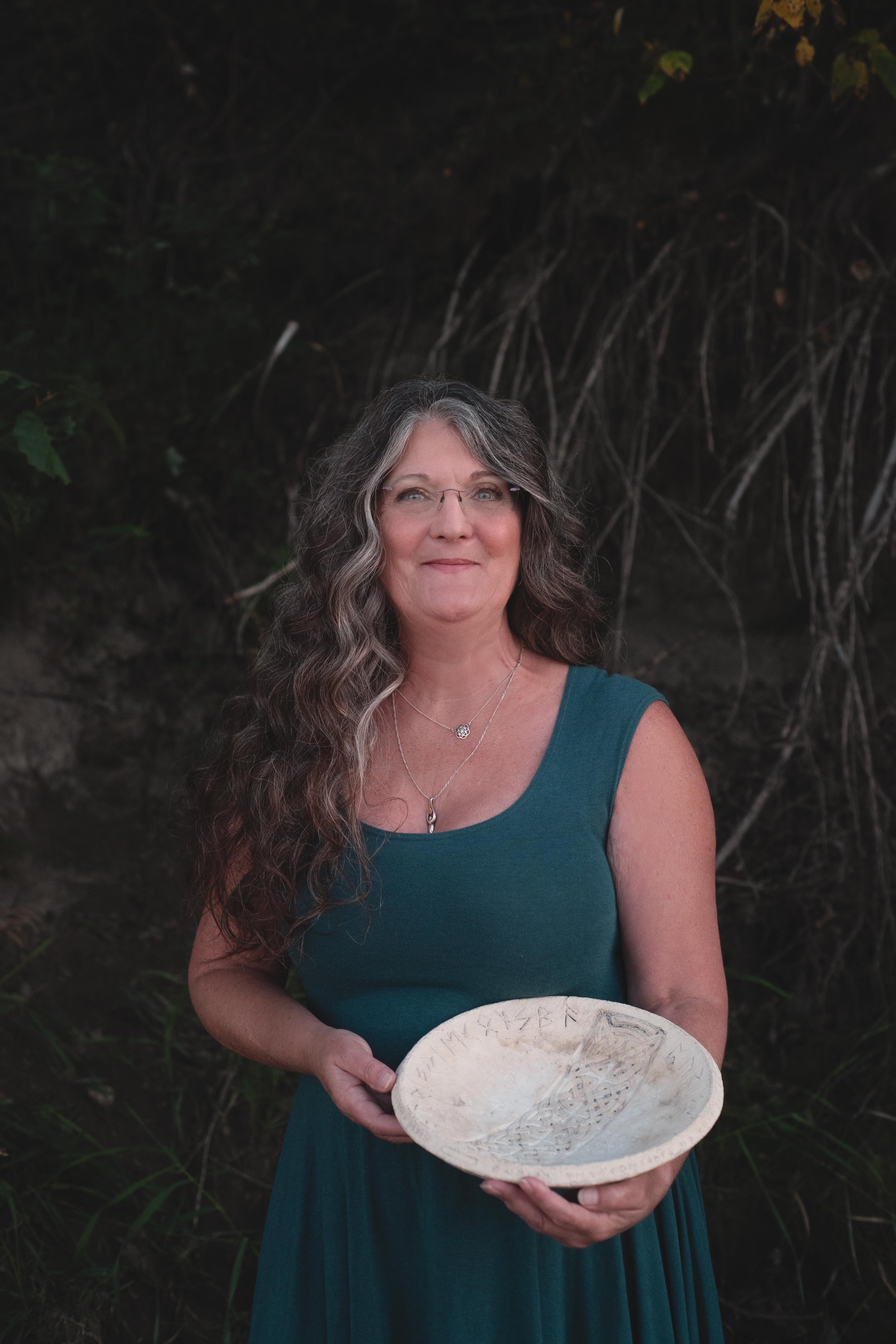 A woman with long, wavy gray hair and glasses, dressed in a teal sleeveless dress, stands outdoors near a dark, leafy background, holding a carved white shell or ceramic artifact with inscriptions.