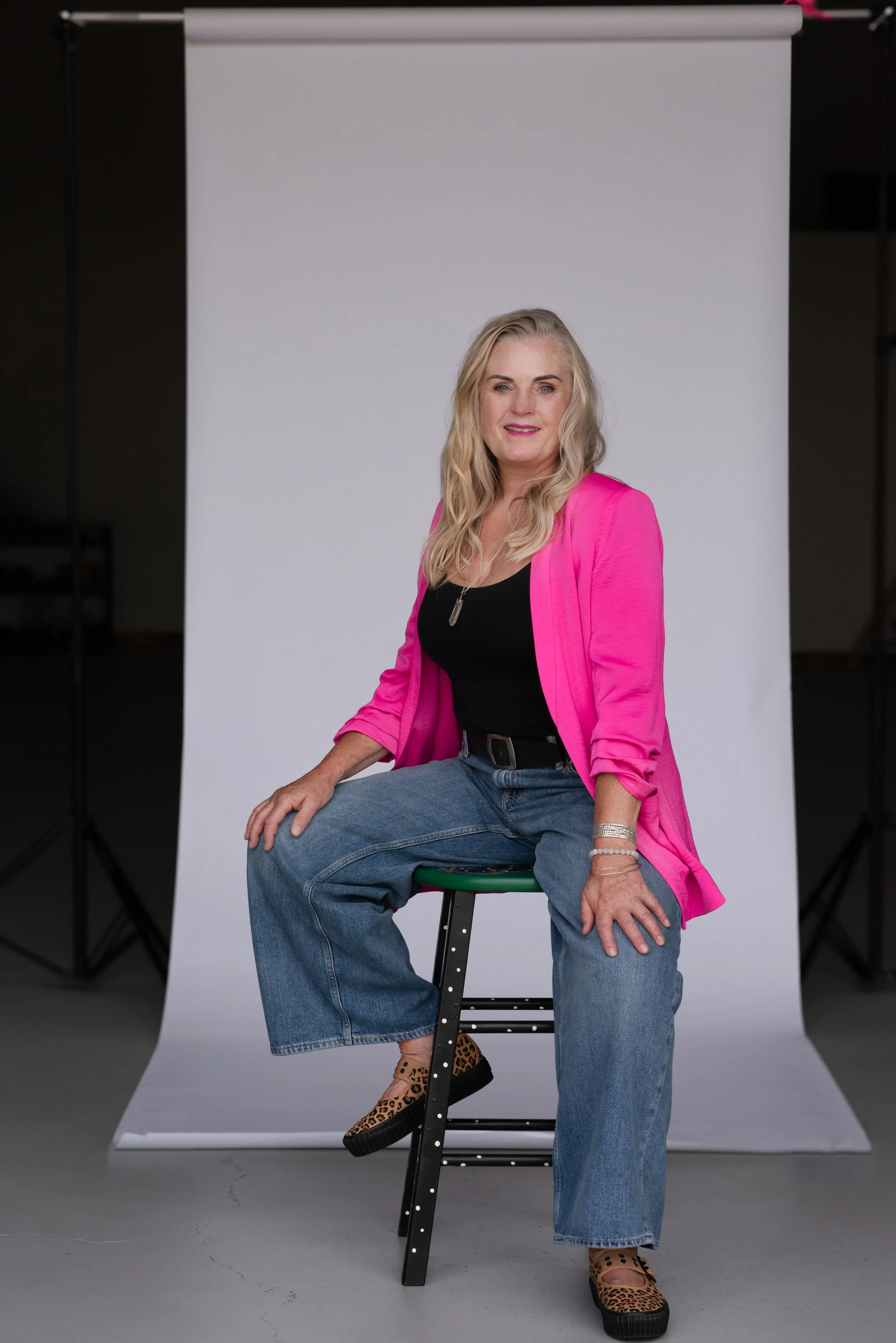 A woman with long blonde hair sitting on a black stool with one leg crossed over the other, wearing a bright pink blazer, black top, oversized jeans, and leopard print shoes, in front of a plain white backdrop.