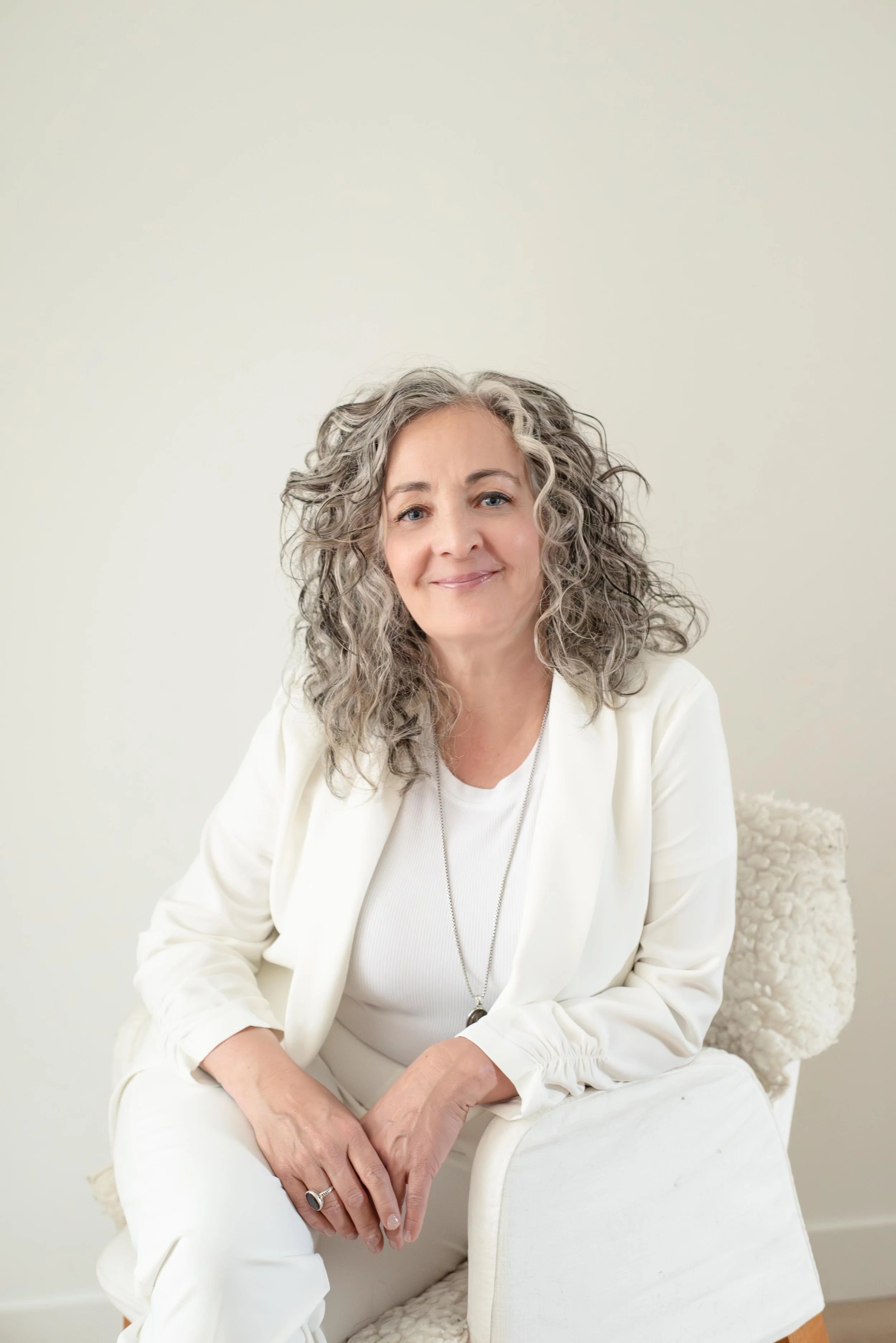 A middle-aged woman with curly gray hair smiling and sitting on a white chair against a plain off-white wall. She is wearing a white jacket, white shirt, and white pants.