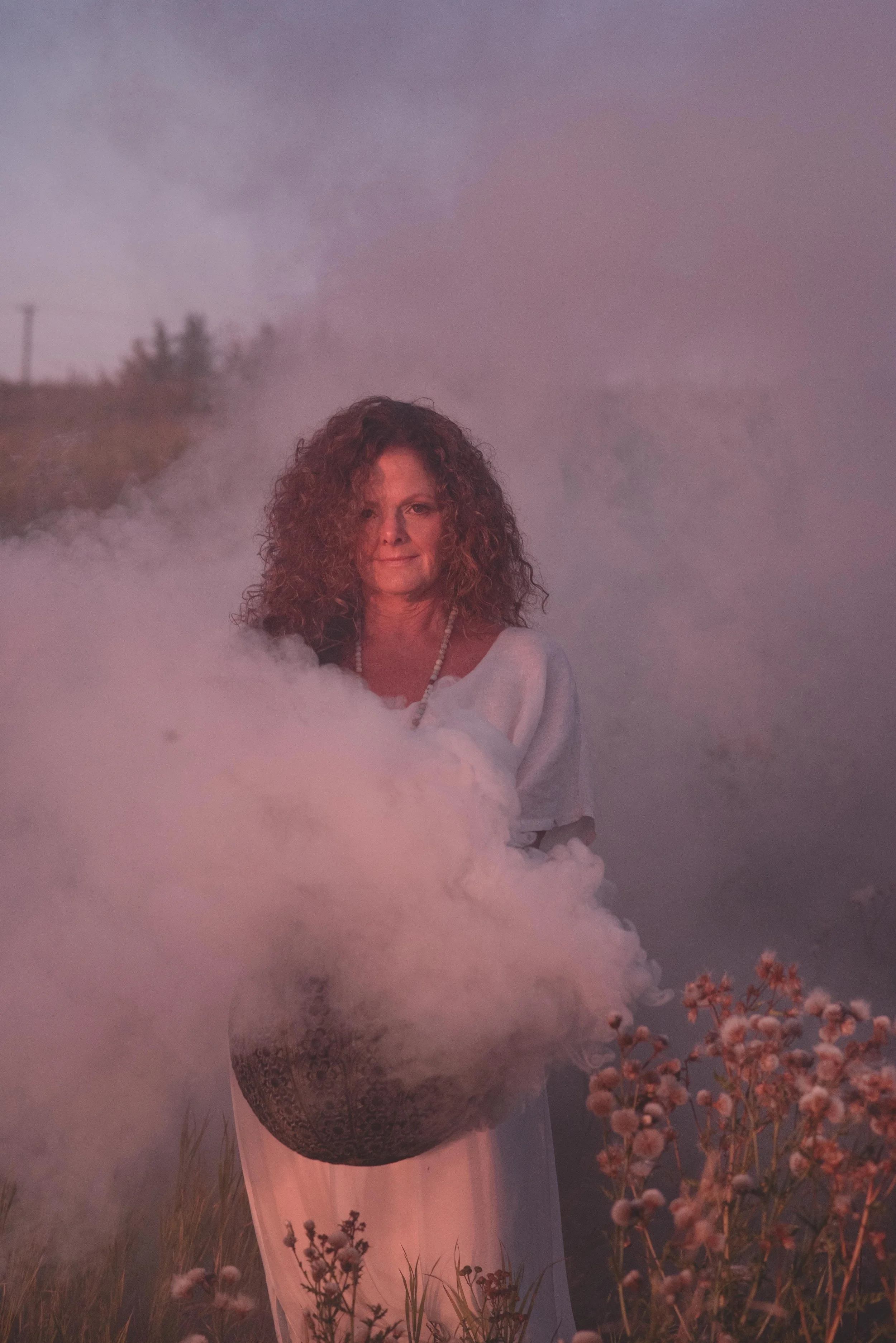 Woman with curly hair standing in a foggy field during sunset, holding flowers.