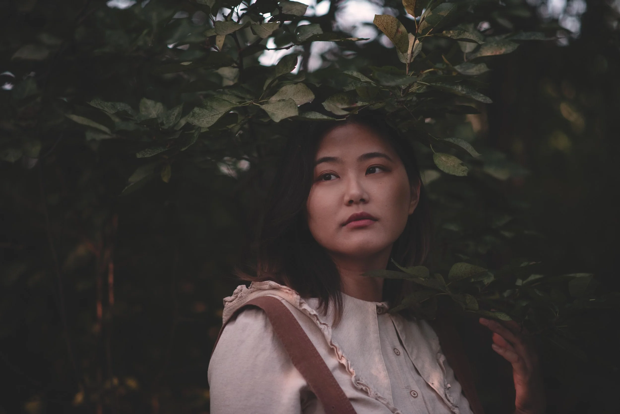 A woman with dark hair and a light-colored blouse stands partially behind dense, dark green leaves and branches, with her face and upper body visible as she gazes into the distance amidst leafy foliage.