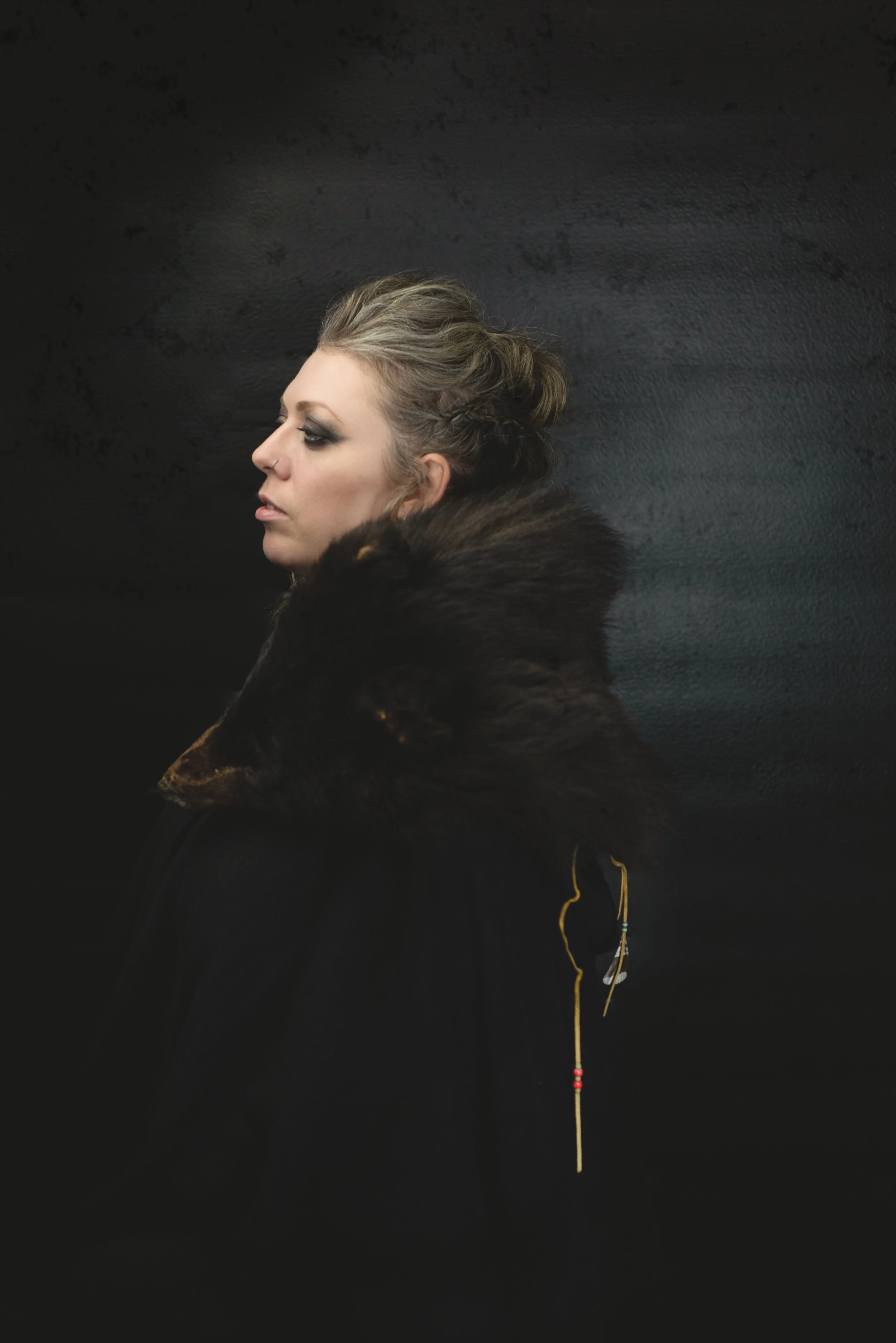 A woman with styled blonde hair and dark eye makeup is wearing a fur coat and looking to the side against a black background.