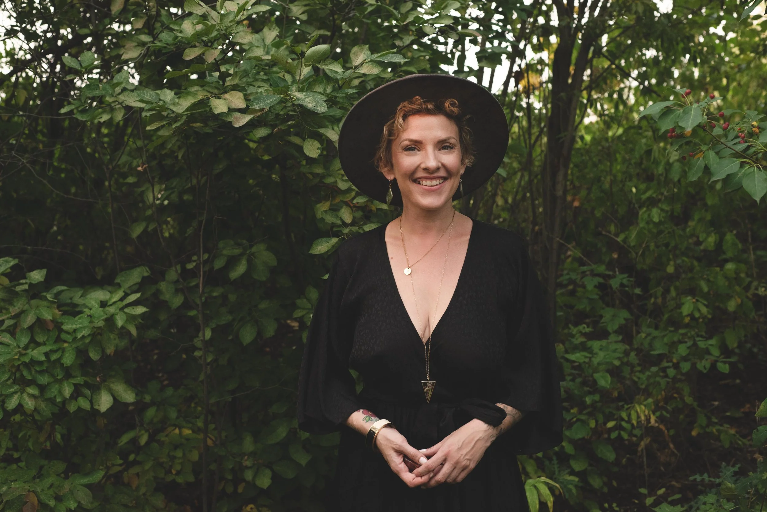 A woman smiling outdoors standing in front of green leafy bushes. She has short curly hair and is wearing a black dress, a wide-brimmed black hat, and jewelry including earrings, necklaces, and bracelets.