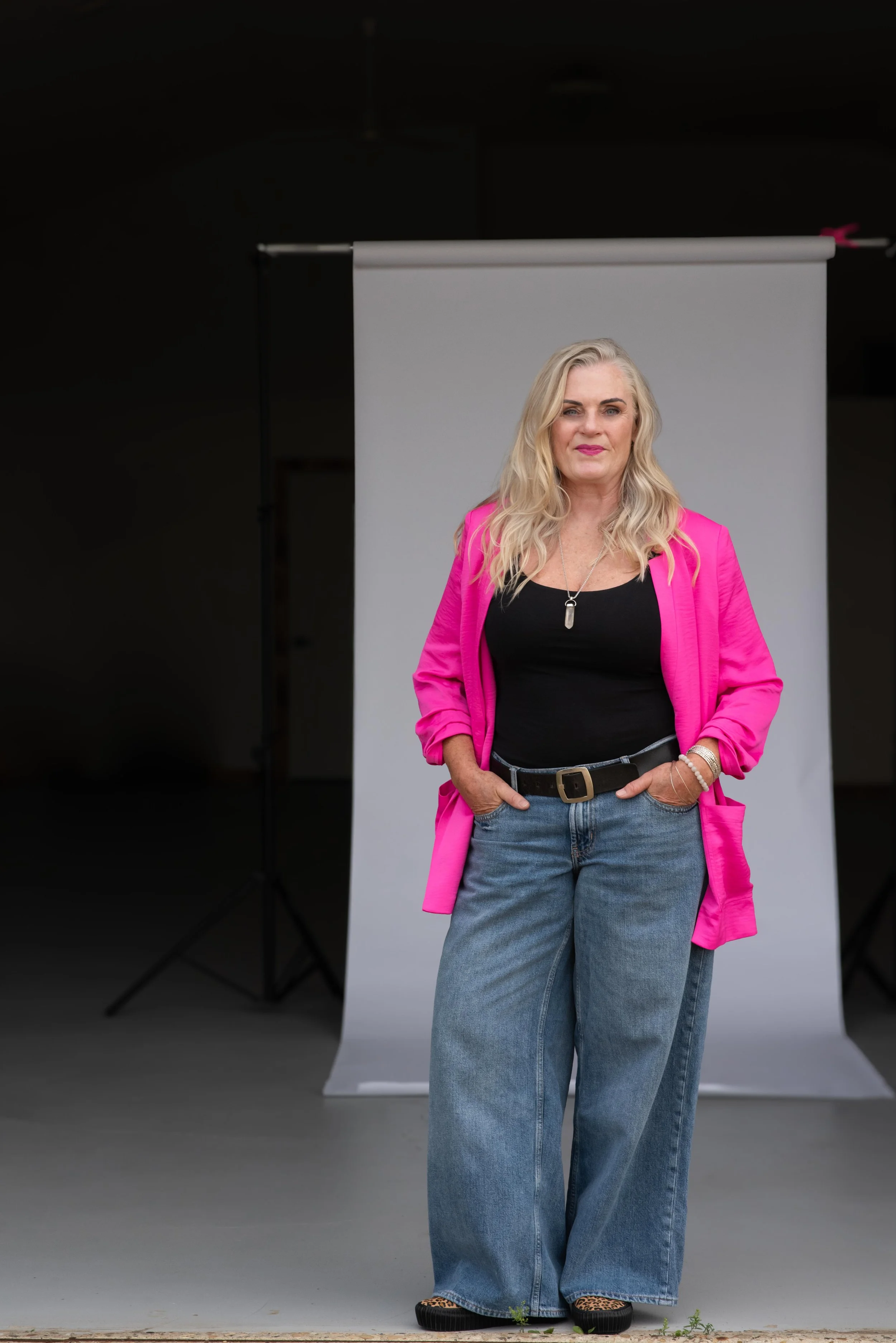 A woman with long blonde hair wearing a pink blazer, black top, blue jeans, leopard print shoes, silver jewelry, and a black belt posing in front of a gray backdrop in a photography studio.