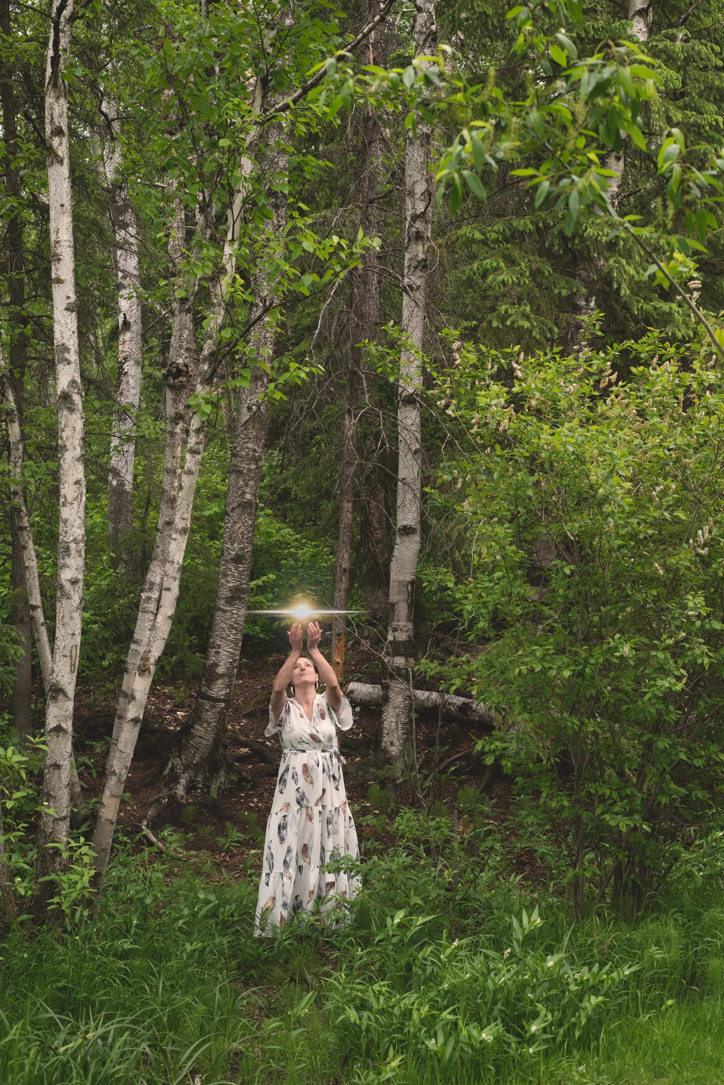 A woman in a white dress with leaf patterns, standing in a green forest, reaching upward towards a glowing light or orb above her hands.