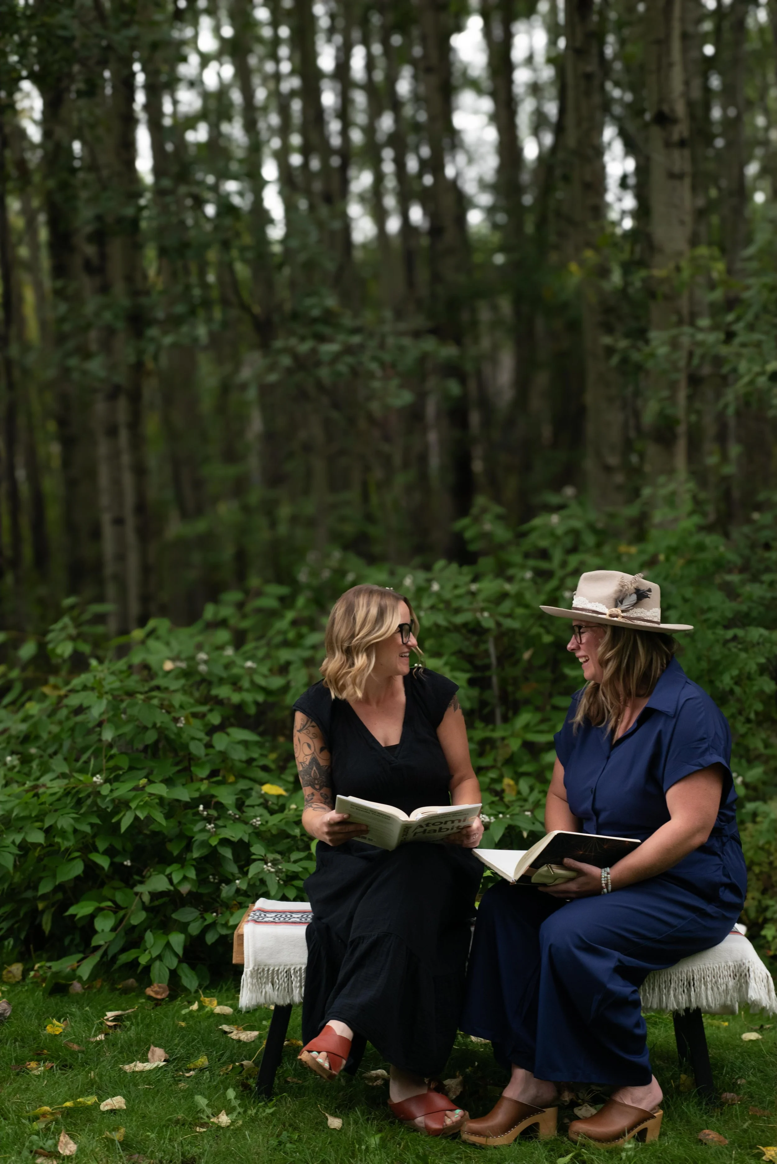 Two women sitting outdoors on a bench, reading books together in a forest setting, one wearing a large straw hat, both smiling and engaging with each other.