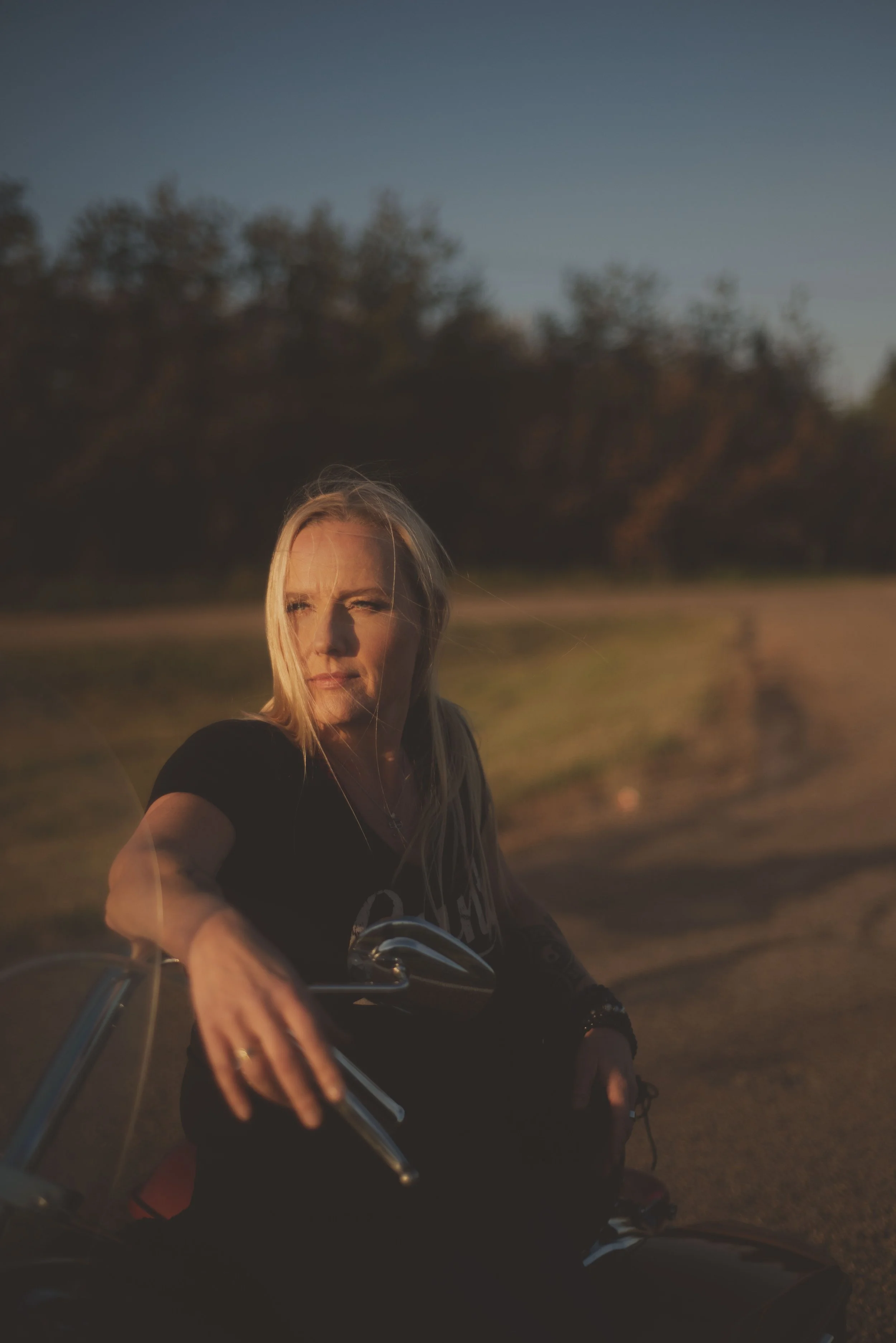 A woman with blonde hair, wearing a black T-shirt, riding a motorcycle outdoors during sunset with trees in the background.