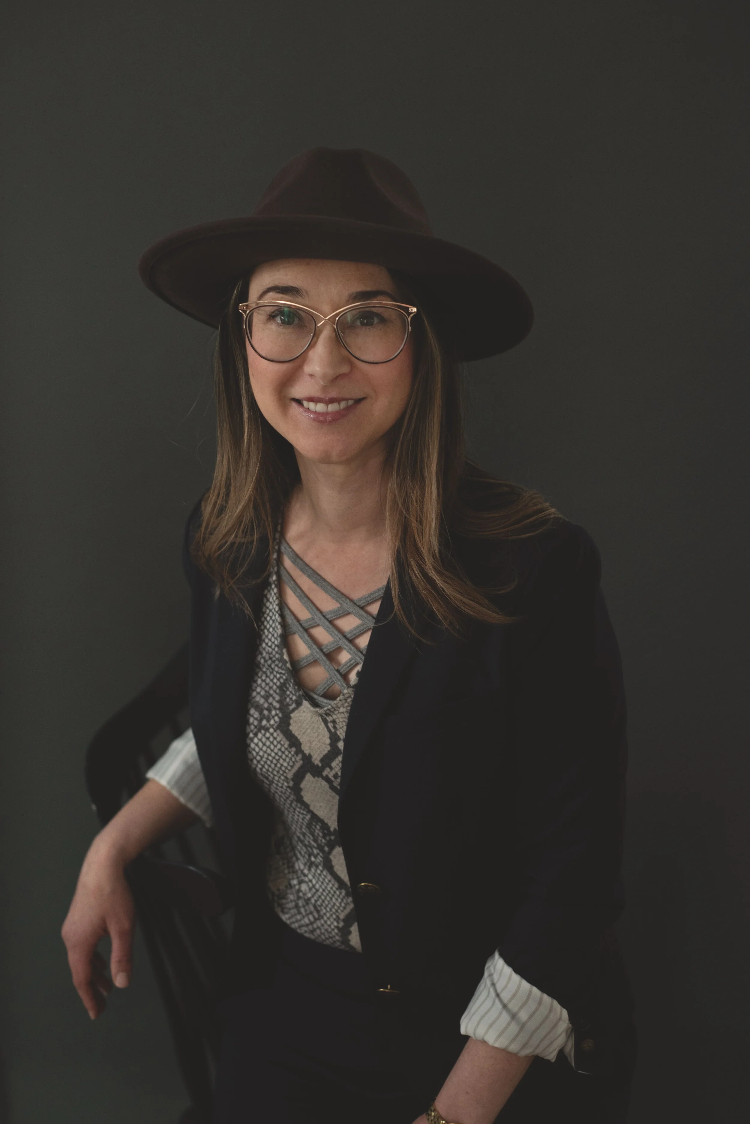 A woman with long, light brown hair, wearing glasses and a wide-brimmed dark hat, is smiling at the camera. She is dressed in a patterned blouse with a crisscross neckline, a black blazer, and has rolled-up sleeves. She is seated in a chair, holding 