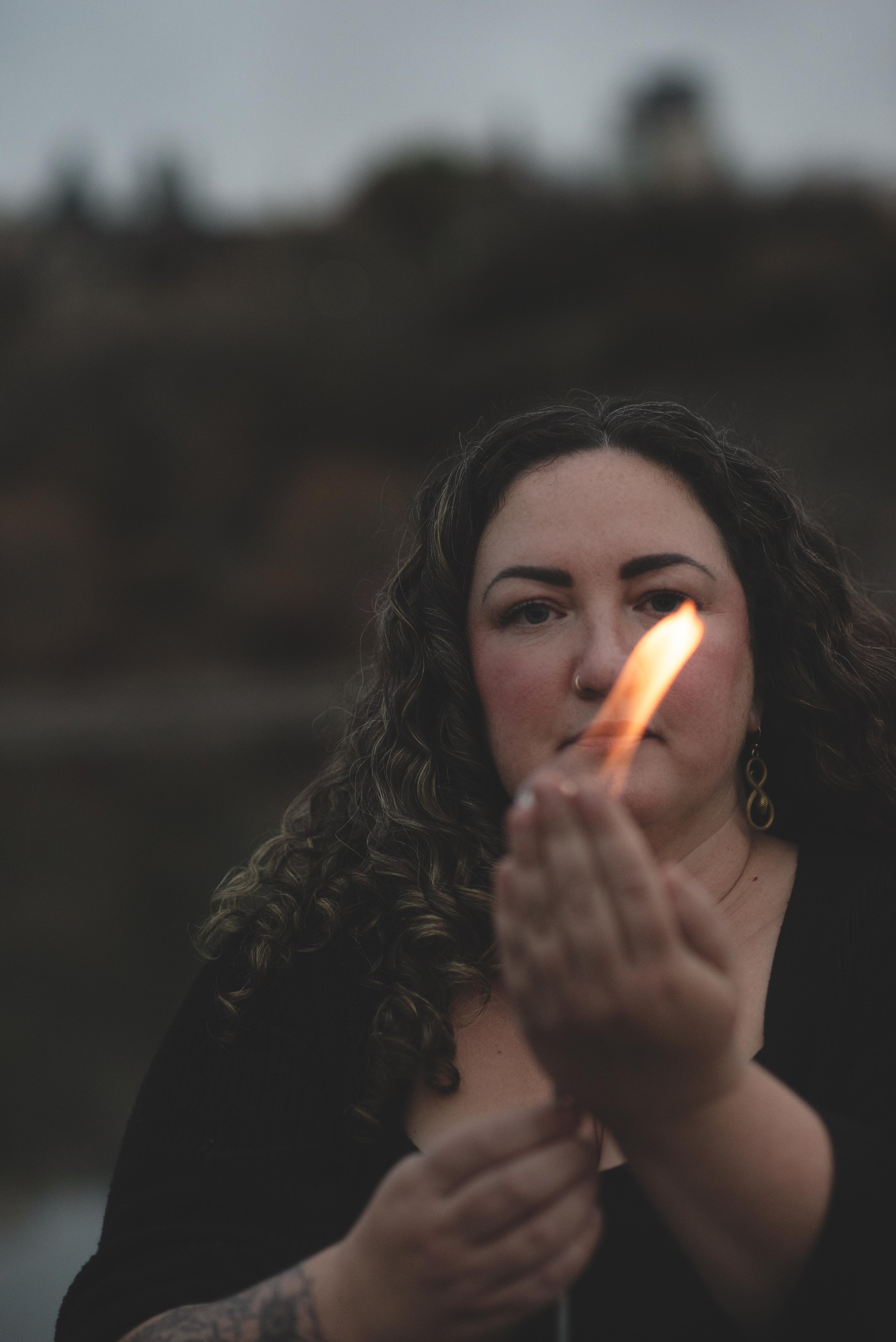 A woman with curly hair, wearing earrings, holding a lit matchstick close to her face with a blurred outdoor background.