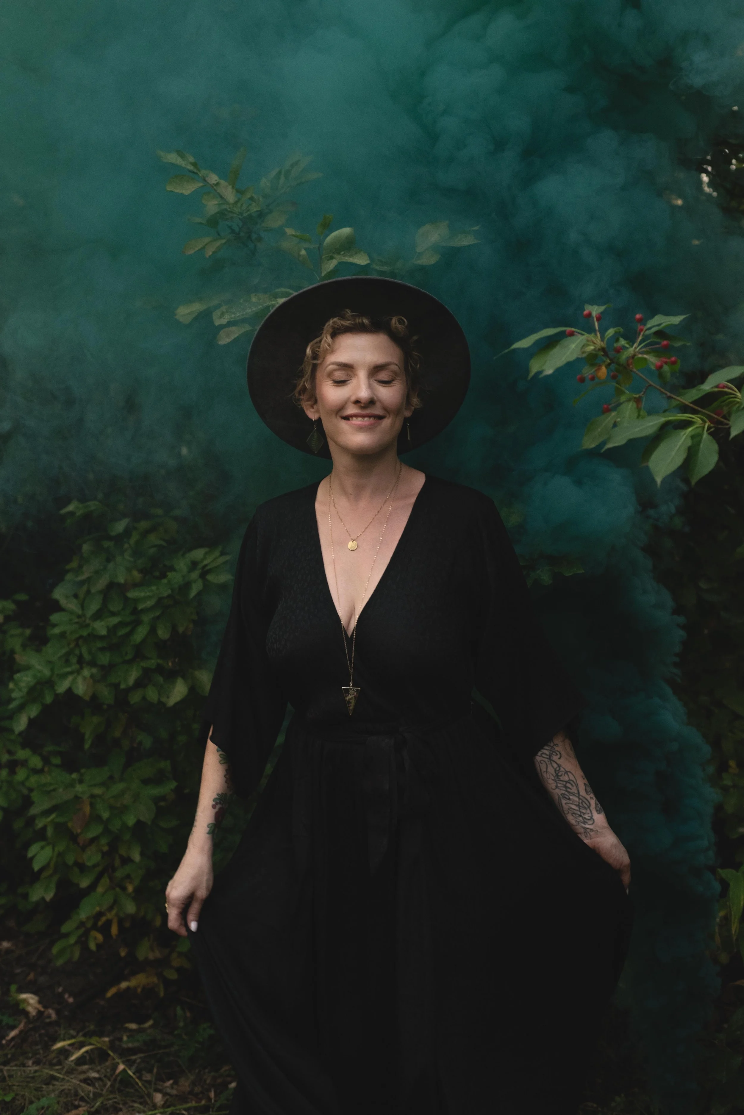 A woman with short, curly blonde hair smiling with eyes closed, wearing a wide-brim black hat, black dress, and layered necklaces, standing in front of green bushes with red berries and a cloud of teal smoke.