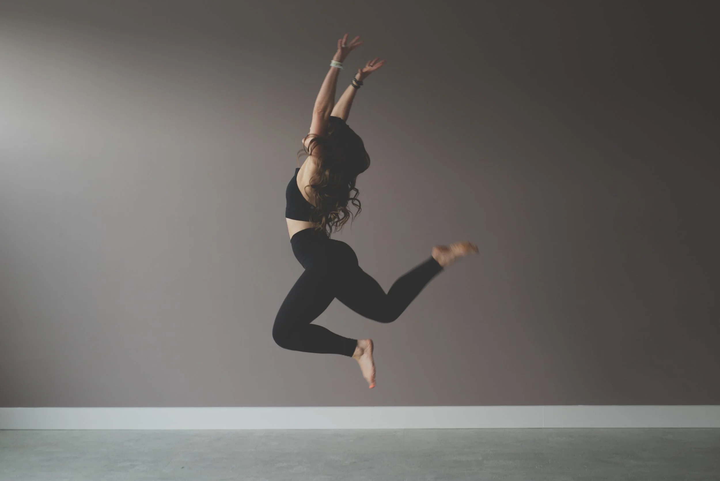 A woman with long hair, wearing a black sports bra and black leggings, is jumping in the air with her arms raised above her head against a plain gray background.