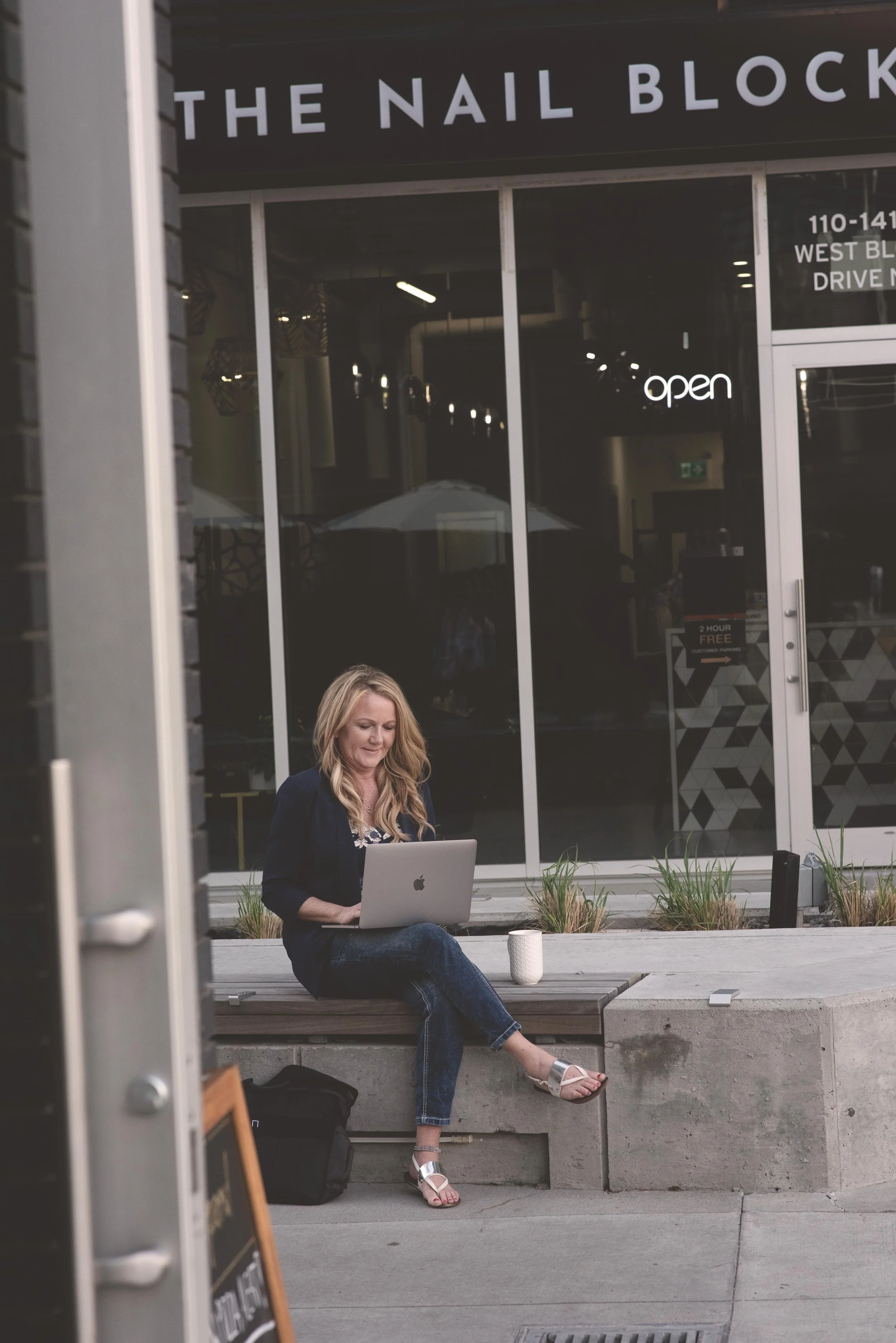 A woman with long blonde hair sitting on a concrete bench outside a cafe, working on a silver MacBook. She is wearing a black top, jeans, and white sandals. There are small plants in pots around her and a white coffee cup on the bench. The cafe behin