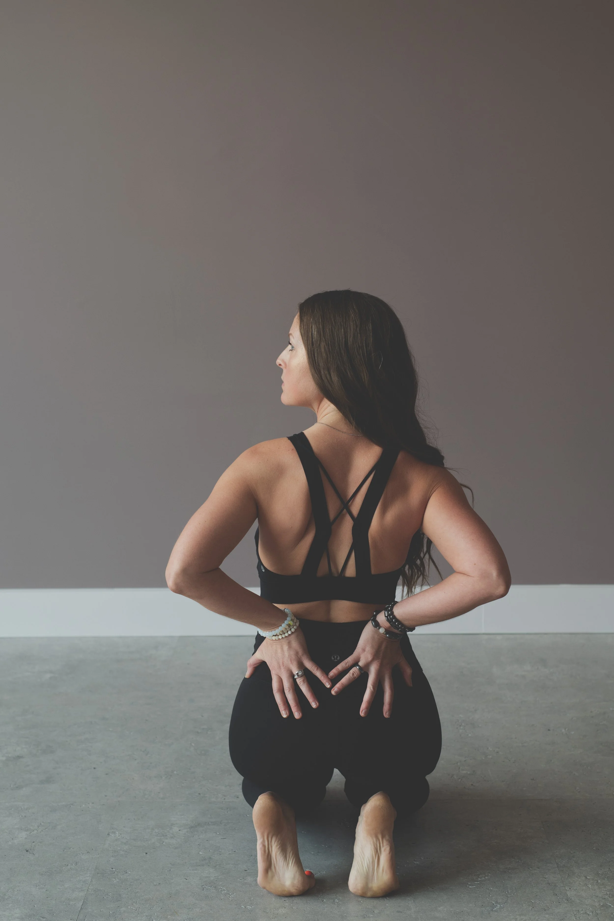Woman in black sports bra and leggings, kneeling on the floor with hands behind her back, facing left against plain gray wall.