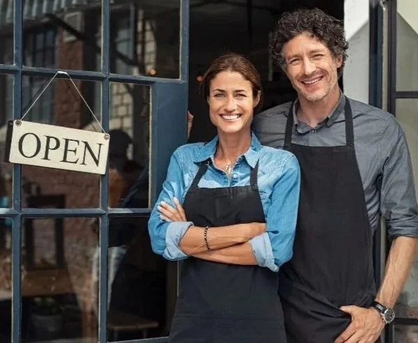 Smiling man and woman restaurant owners standing outside with an open sign in the window.