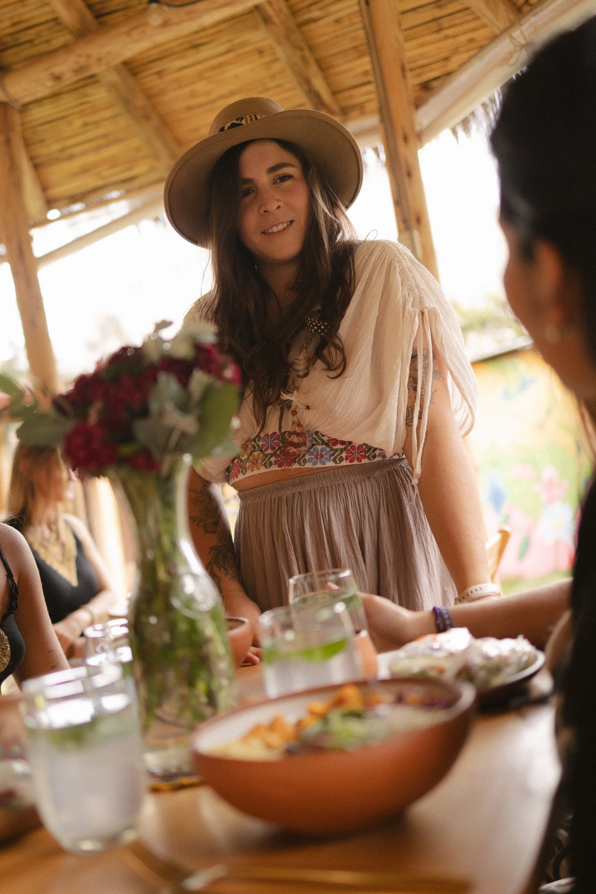 Mujer con sombrero de ala ancha conversando con otra persona en una mesa con comida y flores en un entorno de madera y luz natural.