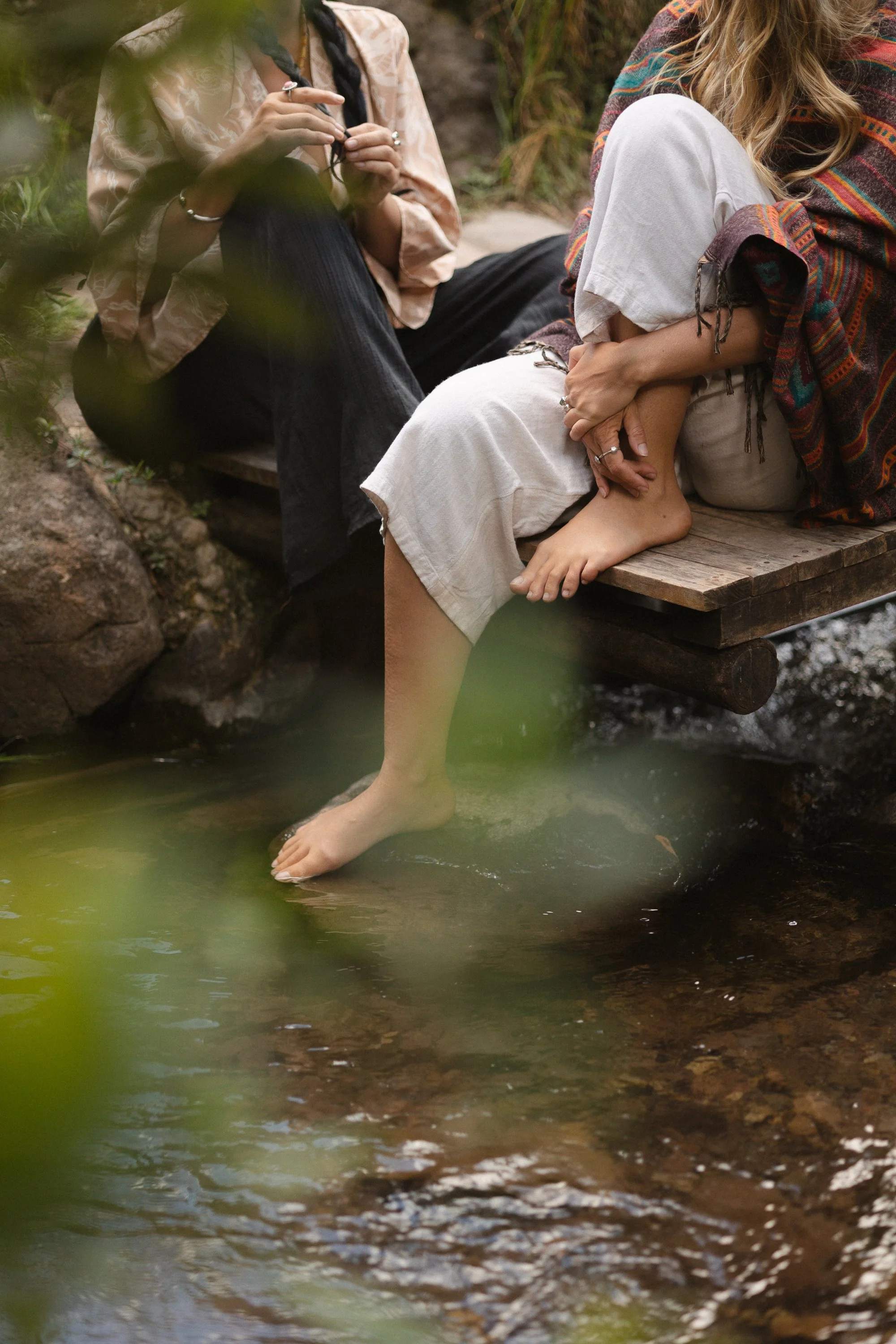 Dos mujeres sentadas en un muelle de madera junto a un río, una de ellas con los pies en el agua y la otra recargada en ella, rodeadas de naturaleza.