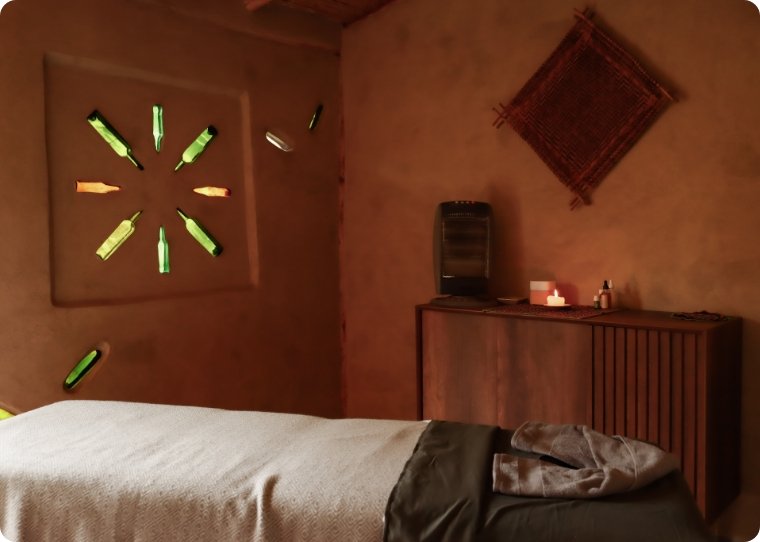 Habitación con paredes de barro o adobe decorada con un tapiz en la pared y una estructura de madera. Hay una cama con sábanas beige, una cómoda de madera, una vela encendida y algunos frascos o pequeños objetos sobre la cómoda. En la ventana hay botellas decorativas o de colores.