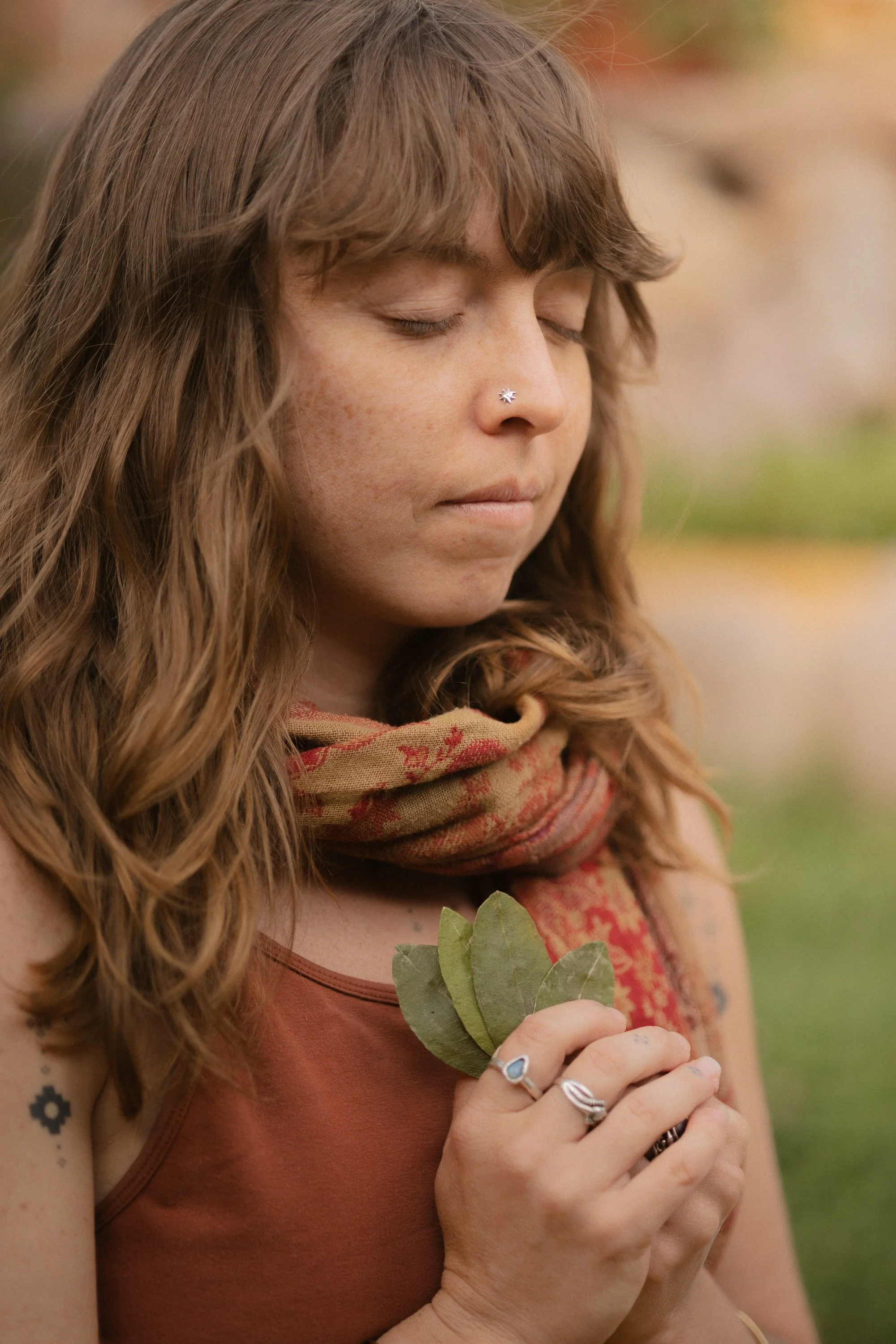 Mujer con ojos cerrados sosteniendo hojas verdes y un pequeño ramo, con tatuajes en el brazo y anillos en los dedos, en un entorno natural.