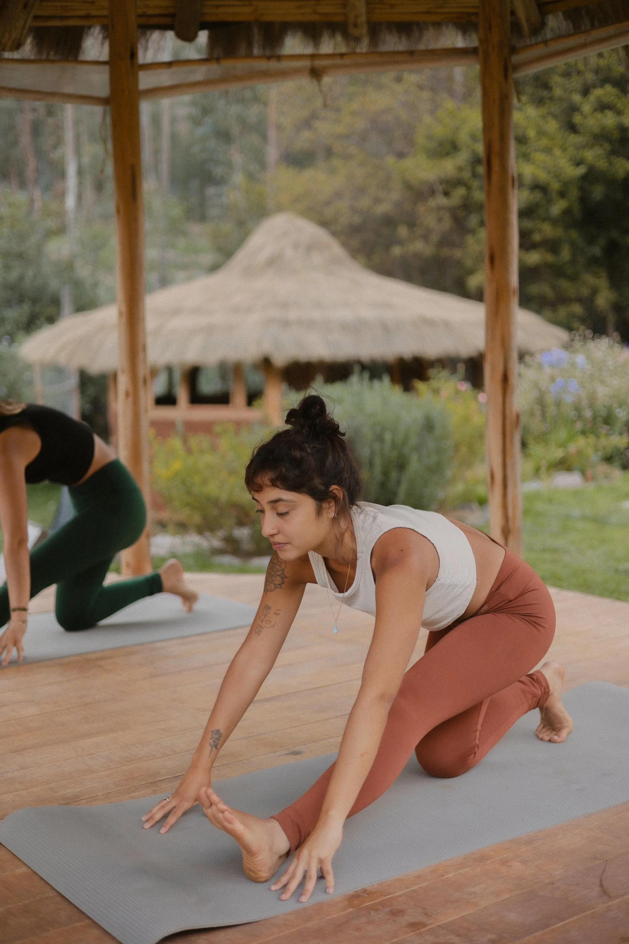 Una mujer practicando yoga en un entorno al aire libre con una estructura de madera y un techo de paja, rodeada de vegetación y un fondo de árboles.