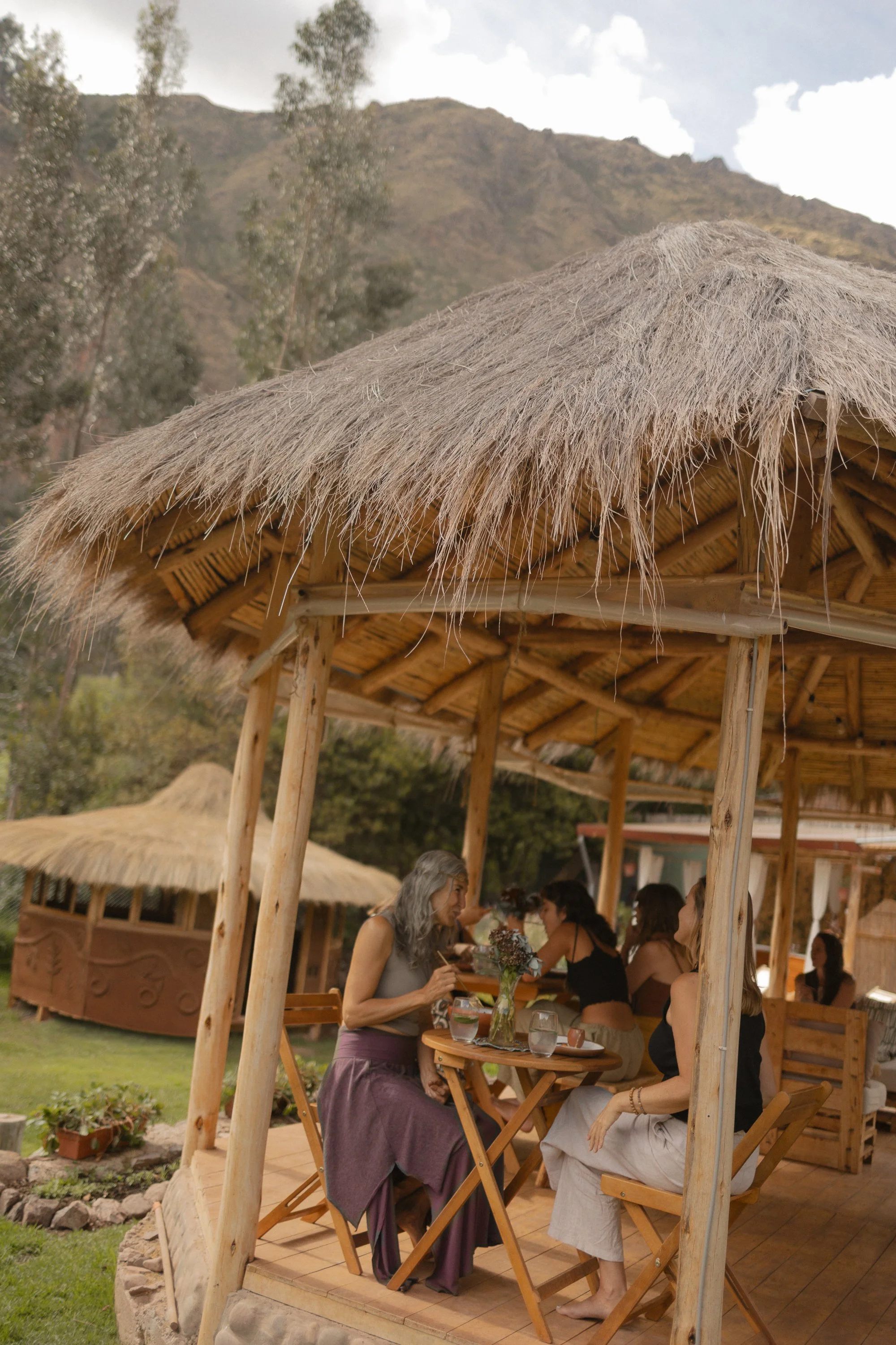 Varias personas sentadas en una mesa en una estructura de madera con techo de paja, en un entorno natural con montañas y vegetación.