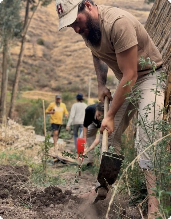 Un hombre con gorra y camiseta marrón cava en una huerta durante el día, con otros en el fondo en un paisaje rural