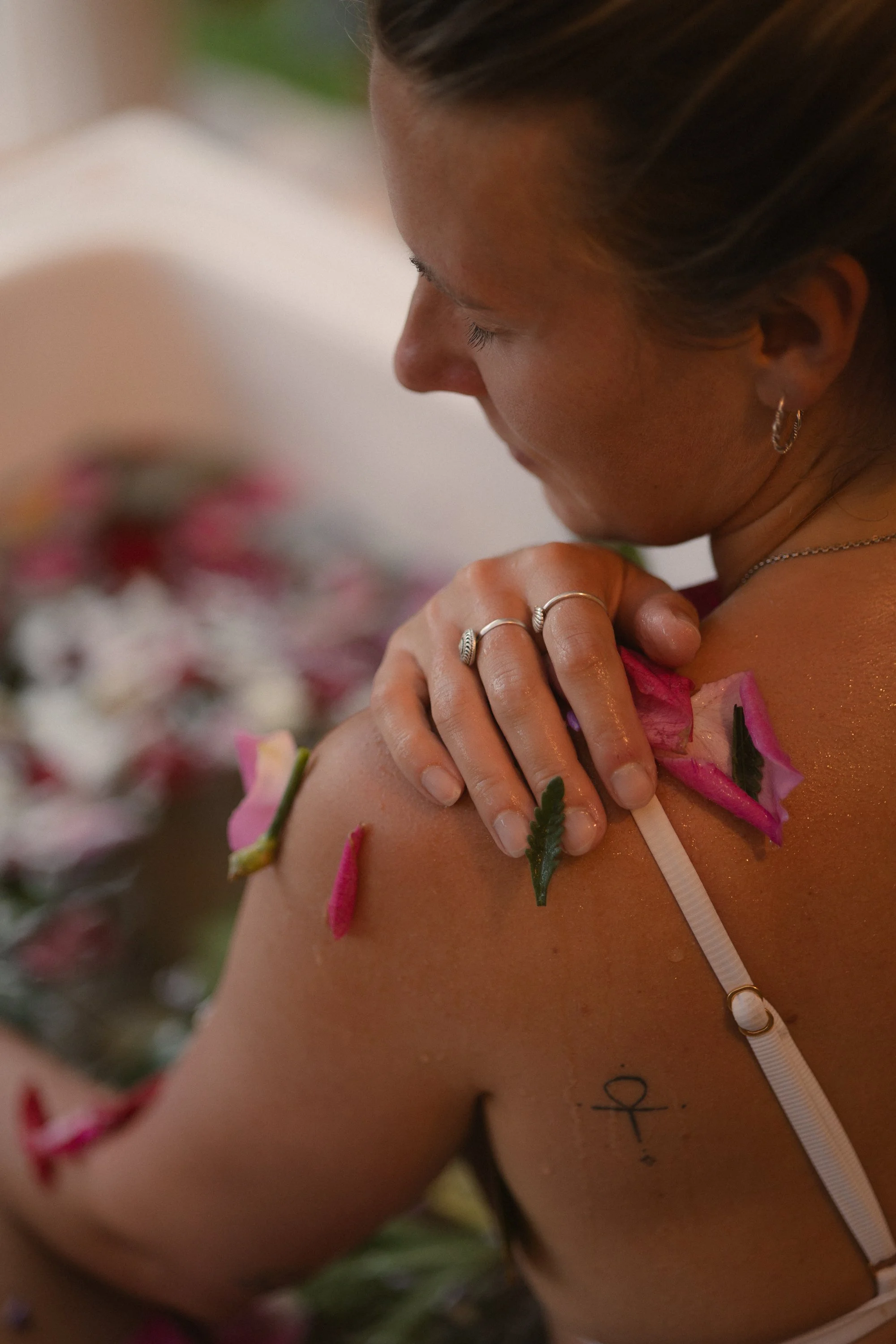 Primer plano de una mujer con tatuaje y joyas, cubriéndose el hombro con flores y hojas, en un ambiente relajado y femenino.