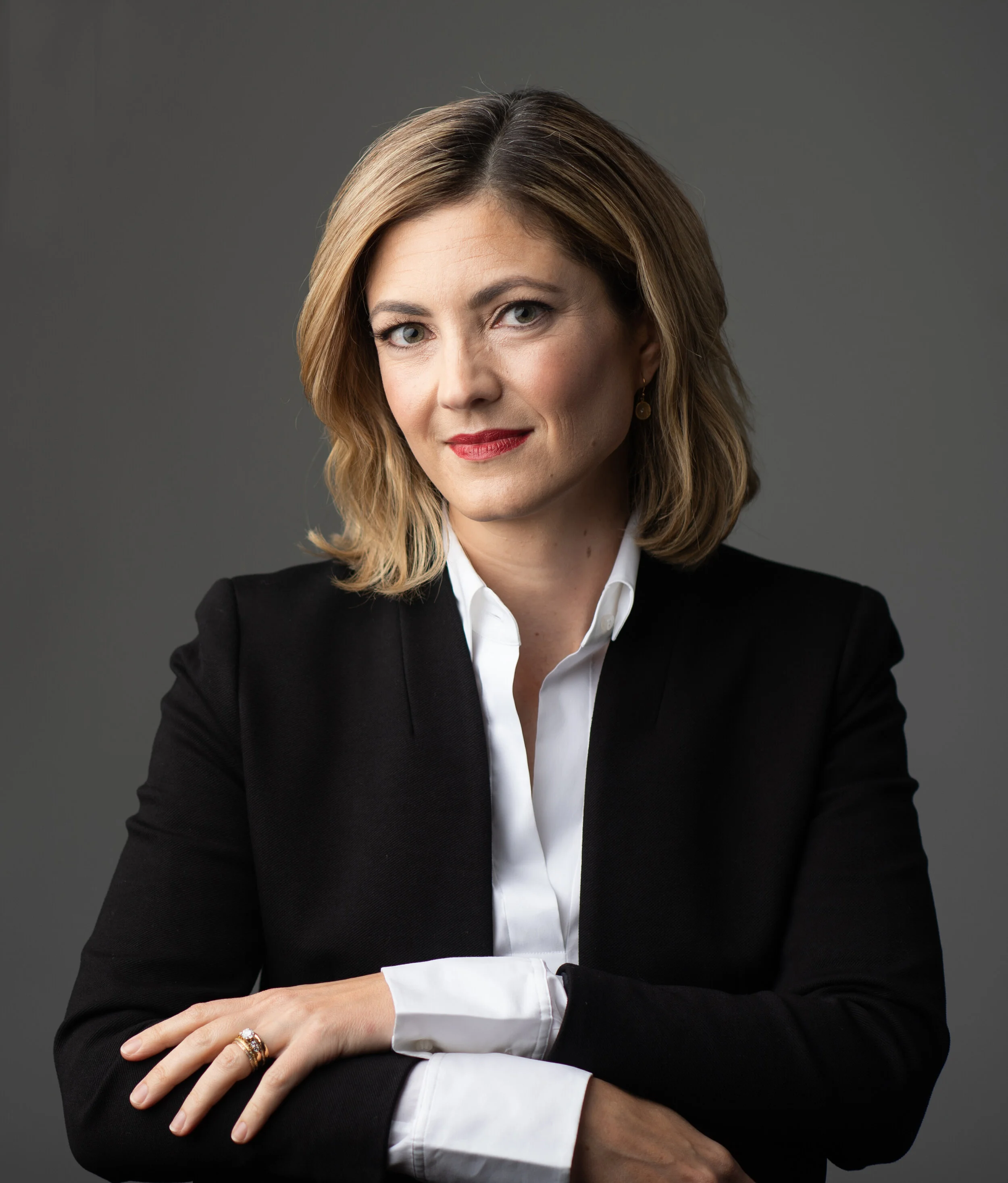 Portrait of a woman with shoulder-length blonde hair, wearing a black blazer and white blouse, against a gray background.