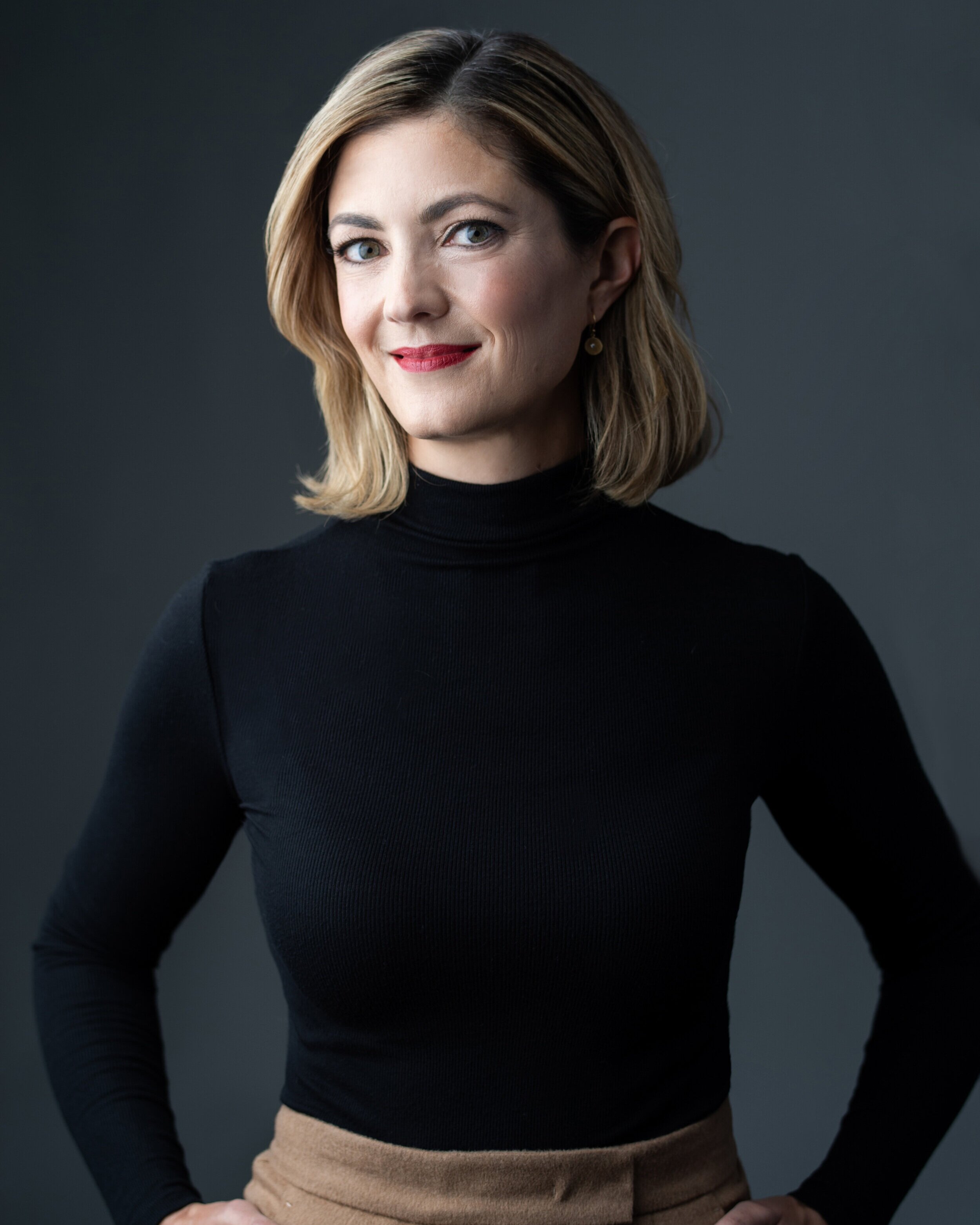 A woman with shoulder-length blonde hair, wearing a black turtleneck and tan pants, smiling at the camera against a dark background.