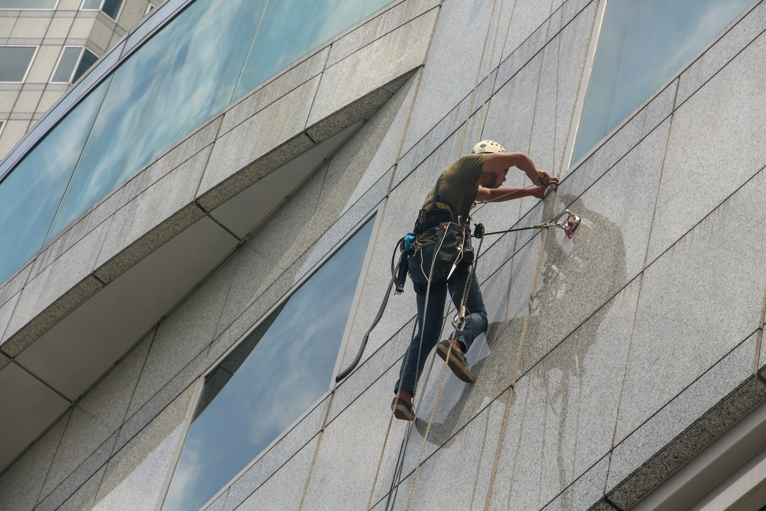 Person wearing safety gear cleaning a glass building facade using ropes.
