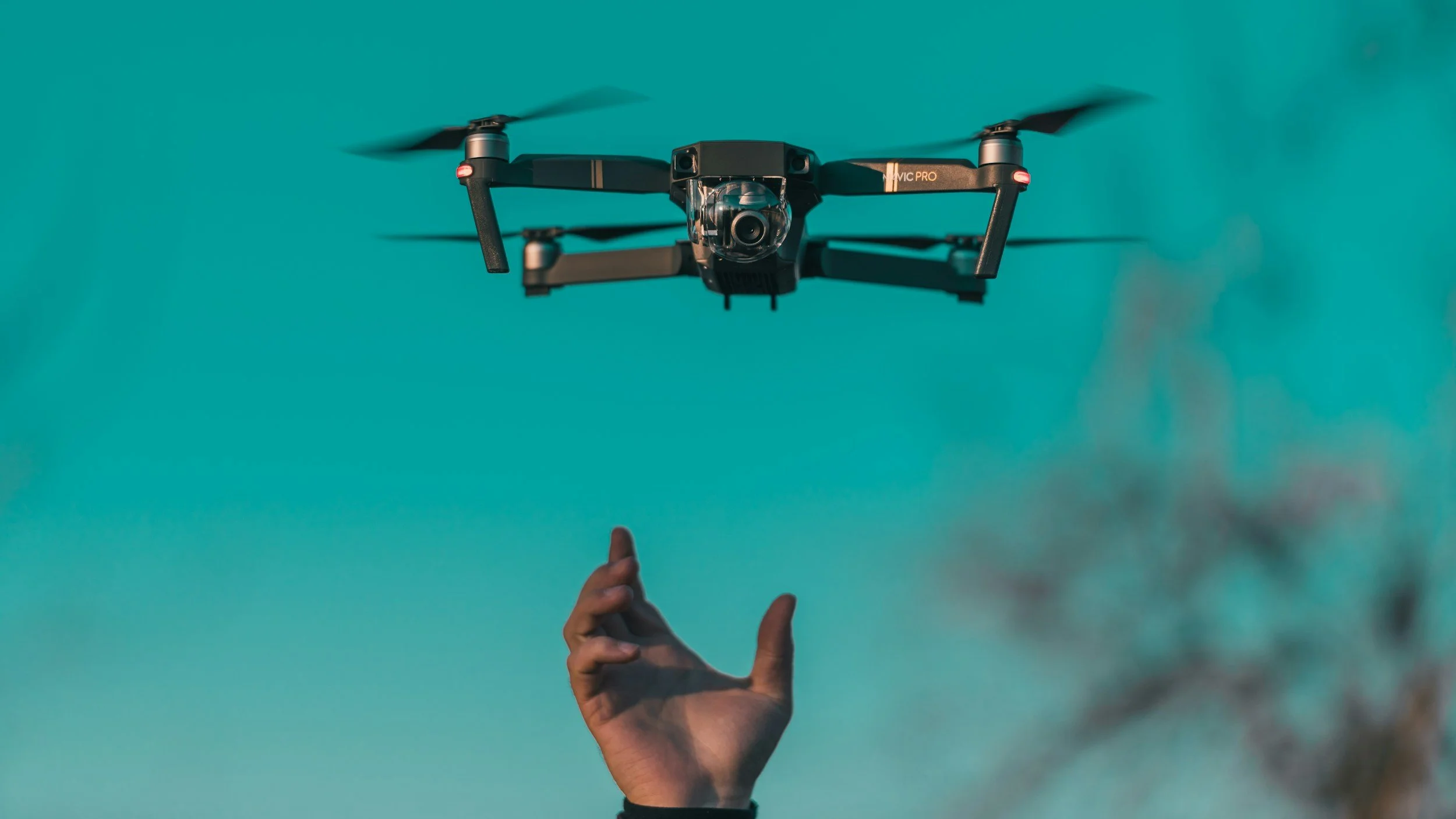 Close-up of a drone in flight with a person's hand reaching up towards it against a clear blue sky.