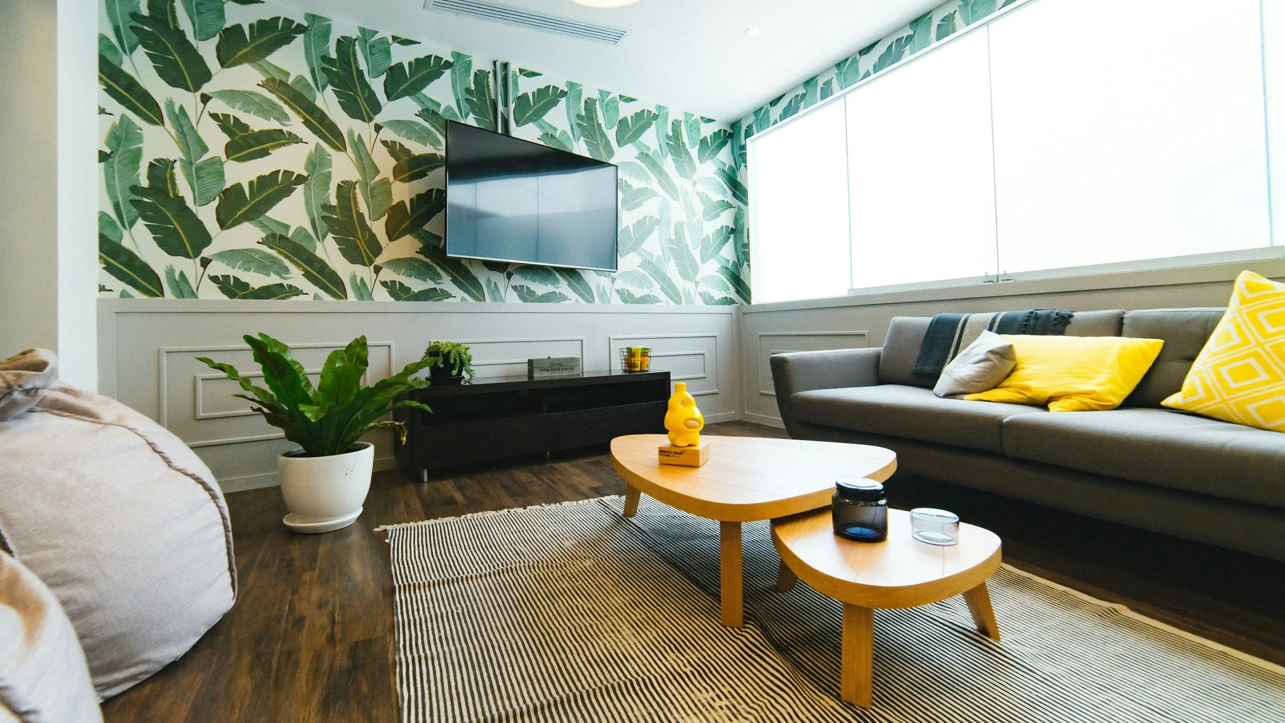 Modern living room with tropical leaf wallpaper, a wall-mounted TV, gray sofa with yellow cushions, wooden coffee table, and a white potted plant on a striped rug.