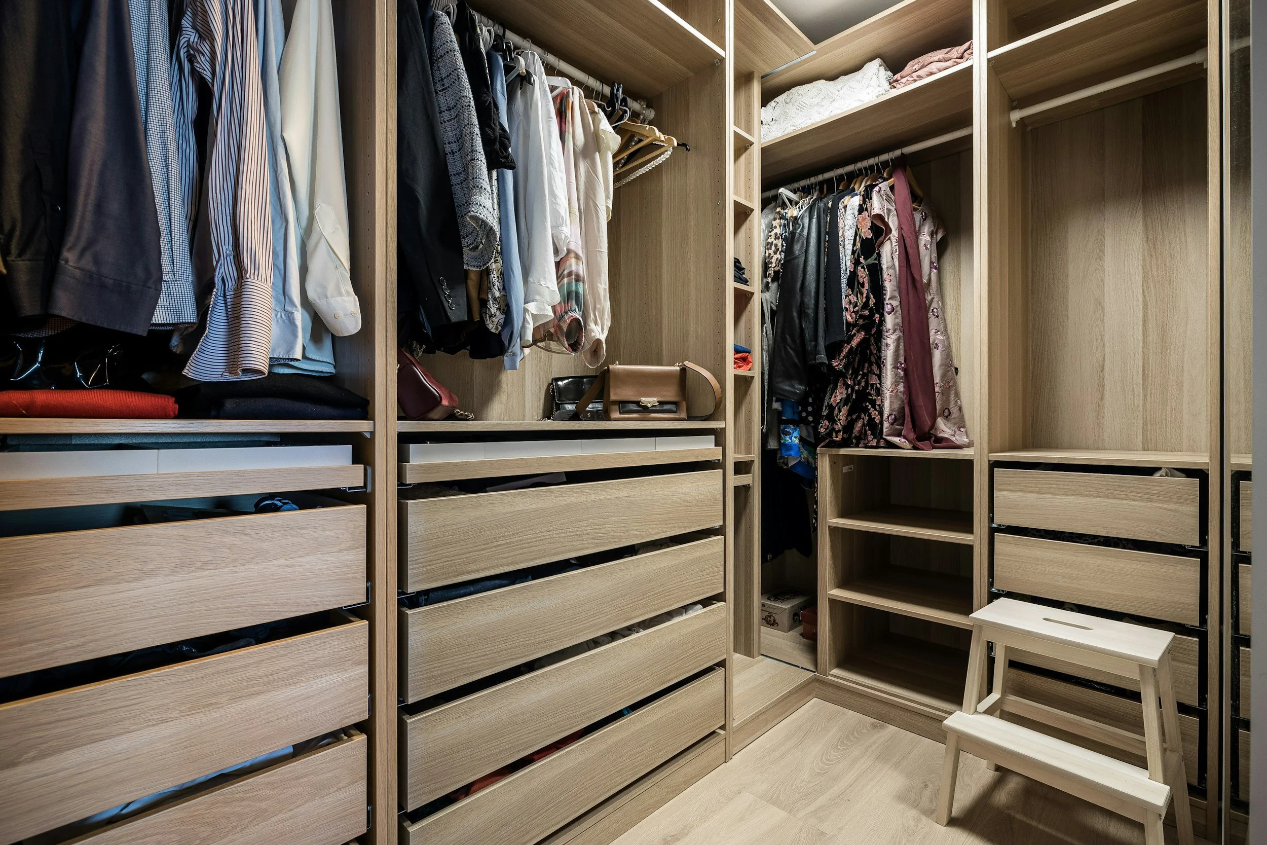 A walk-in closet with wooden shelving and drawers filled with folded clothes, hanging shirts, blouses, jackets, and accessories. A small wooden step stool is in front of the shelves.