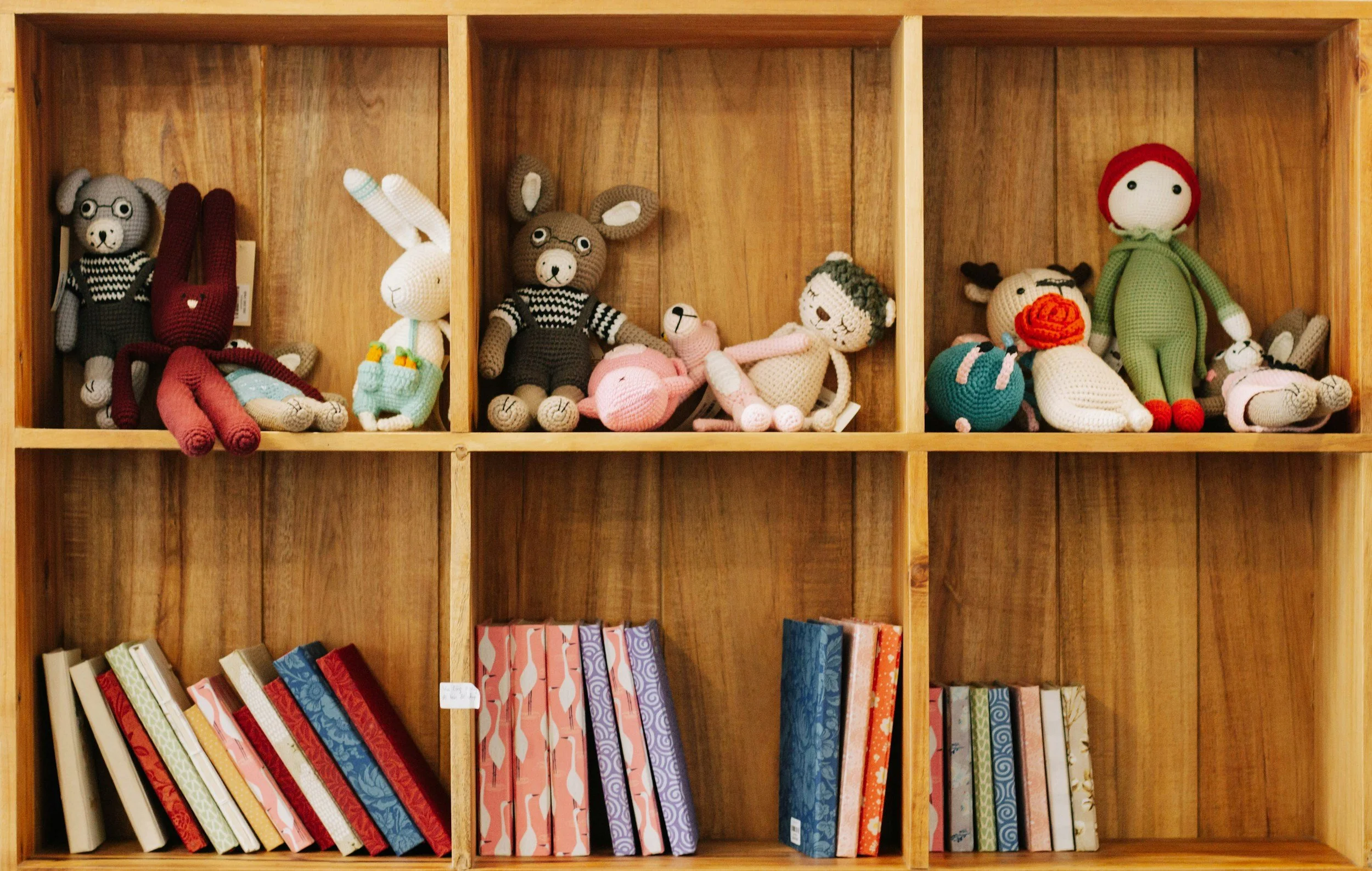 A wooden shelf unit with nine compartments; the top three contain crocheted stuffed animals including rabbits, bears, a mouse, a girl, and a girl with a red hood, with some lying down and others sitting, and the bottom six compartments contain colorf