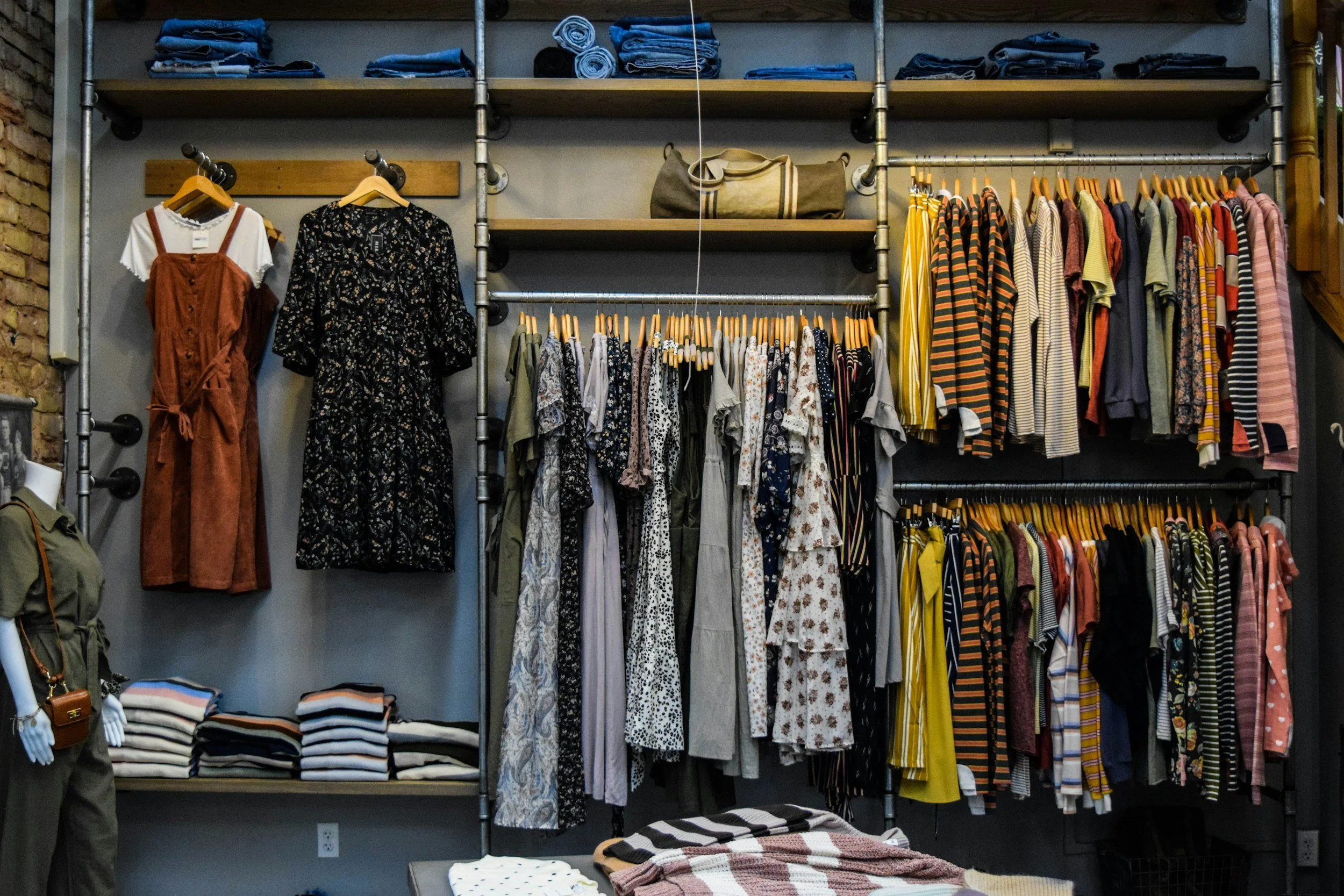 Clothing store display featuring hanging dresses, shirts, and pants, with folded clothes on shelves and bags on upper shelves.