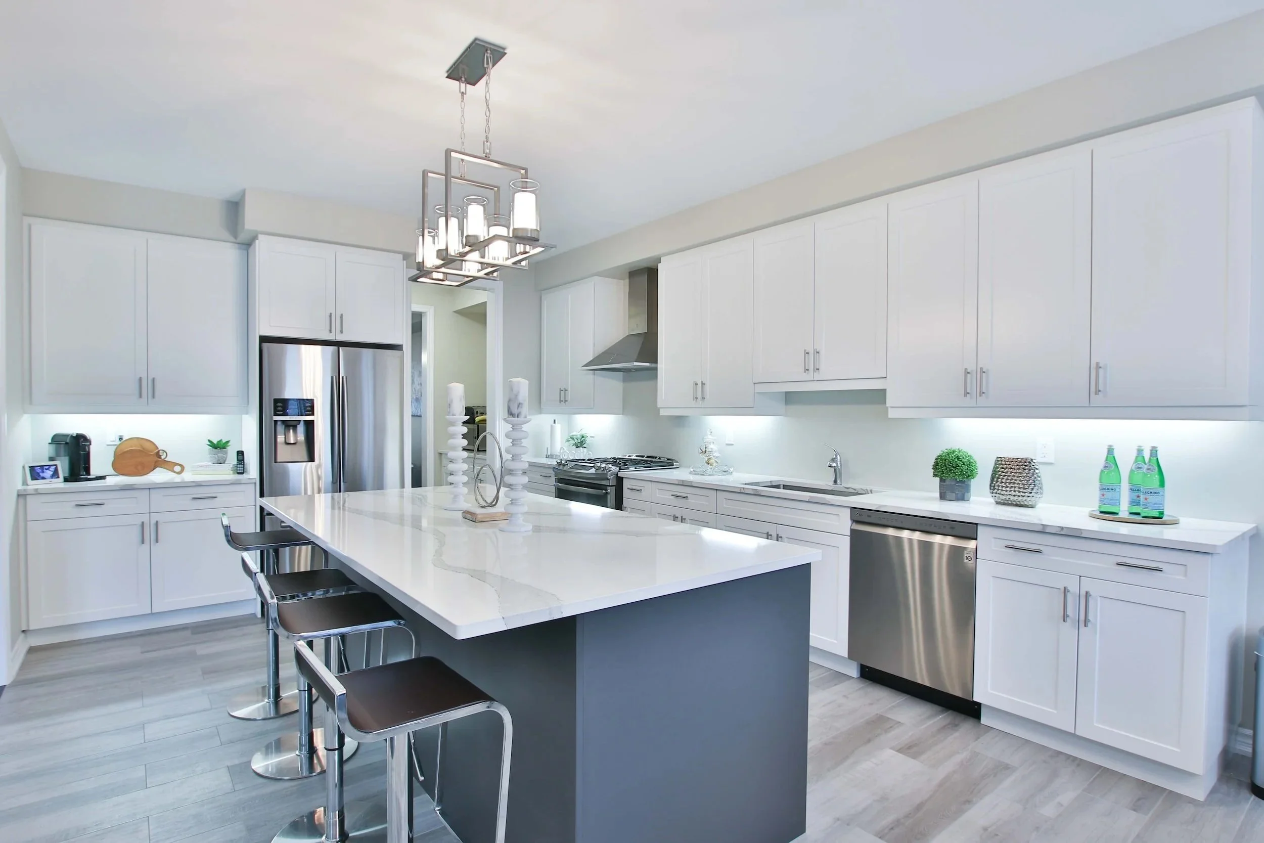 Modern kitchen with white cabinets, stainless steel refrigerator, and a gray island with a white countertop. There are bar stools at the island, decorative candles, plants, and bottles on the counters. A chandelier hangs above the island.