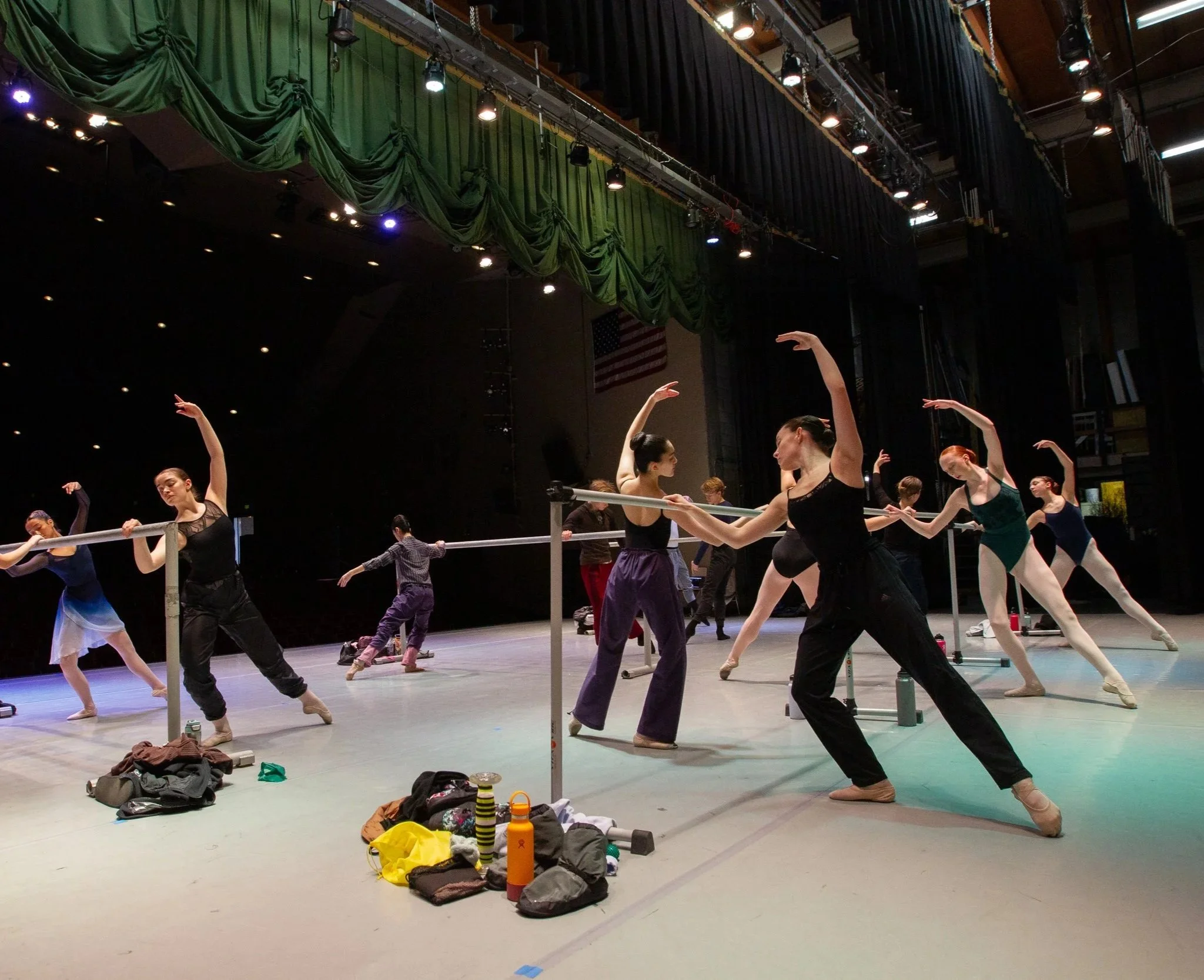 Ballet dancers practicing at a ballet barre in a large theater or rehearsal space. Shoe bags and water bottles are scattered on the floor in the foreground. The stage has green curtains and an American flag hanging in the background.