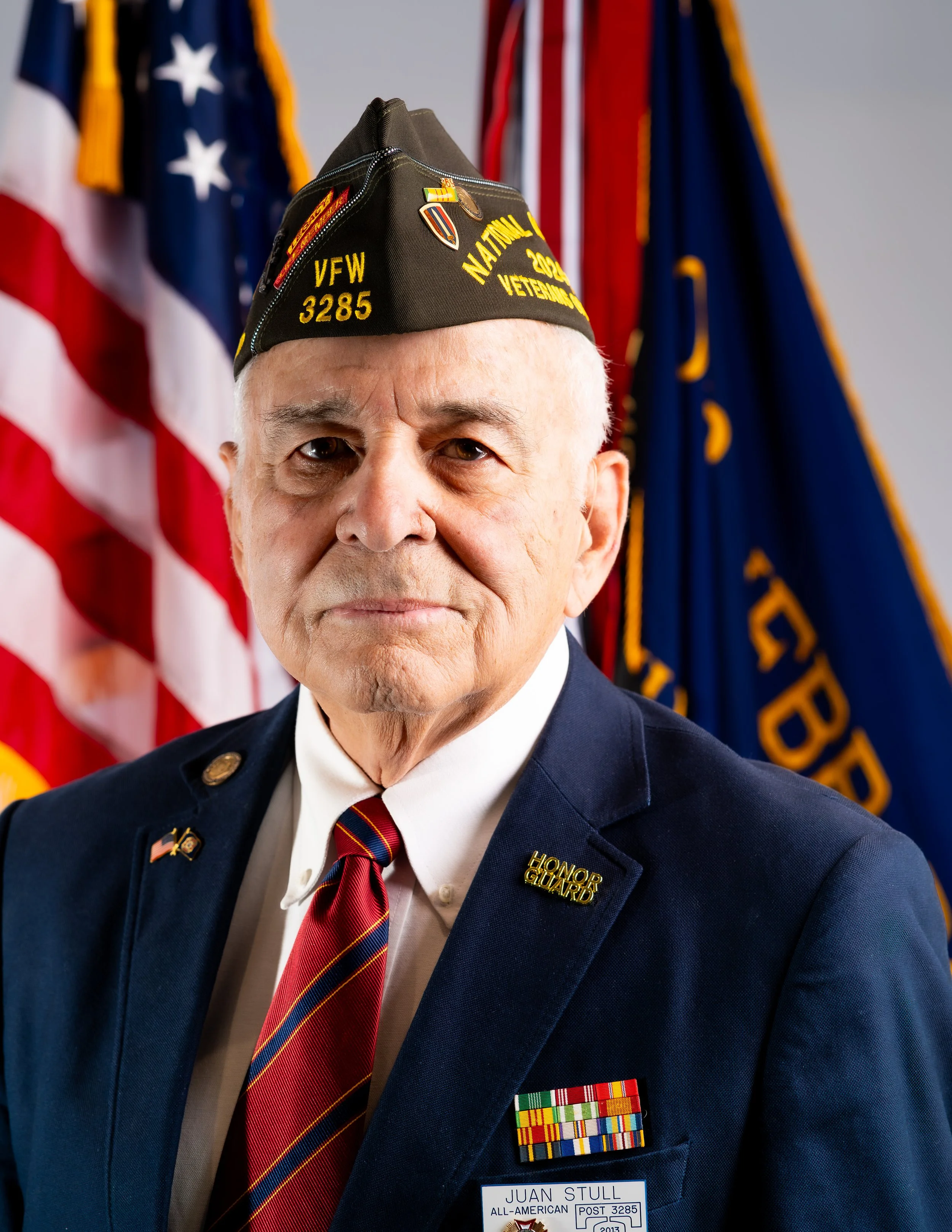 An elderly man in a navy blazer and a red striped tie, wearing a black veterans hat, standing in front of a backdrop of flags.