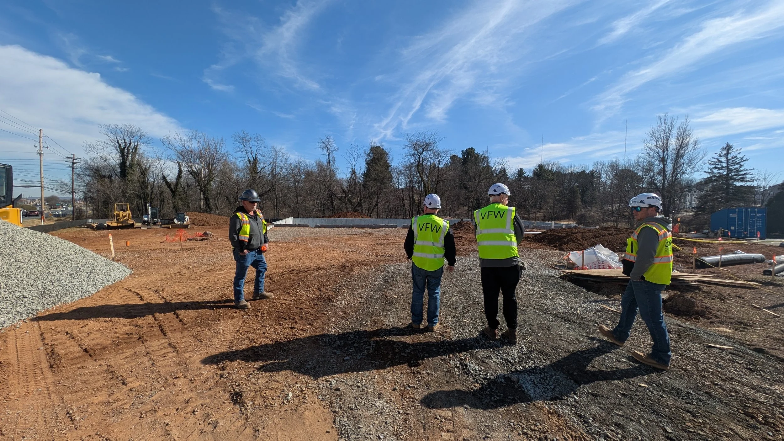 Four construction workers wearing hard hats and reflective vests standing on a construction site with dirt and gravel, with a clear blue sky and trees in the background.