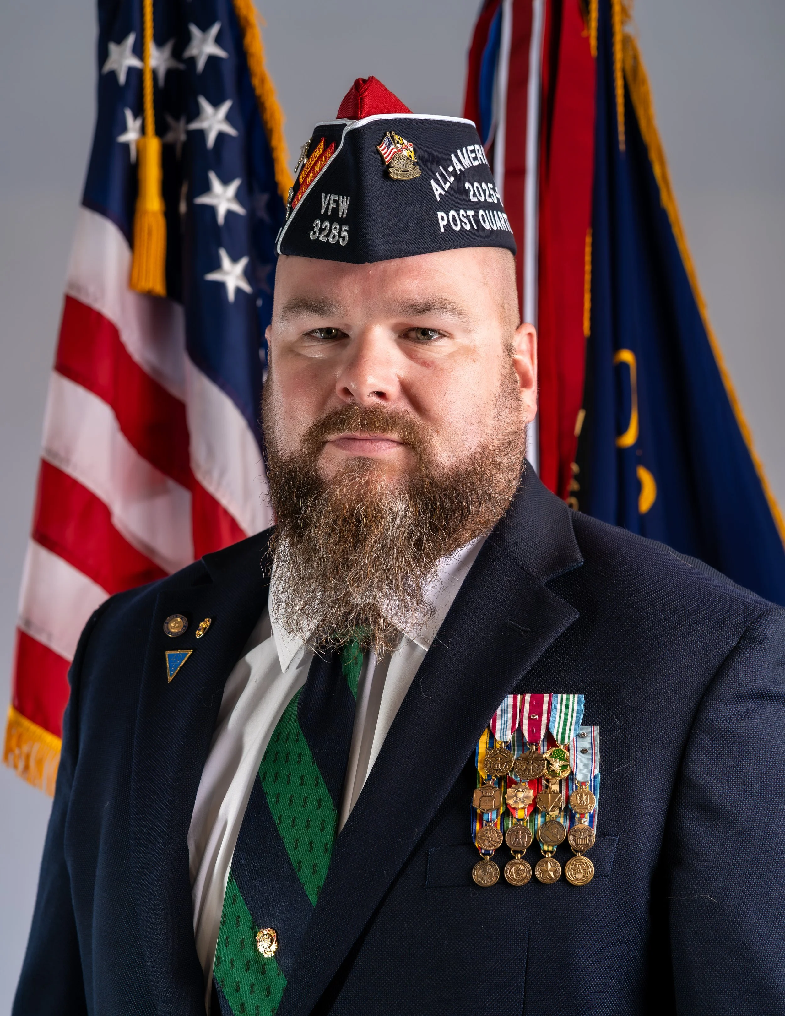 A man wearing a black leather vest with patches, an orange collared shirt, and a black for pride hat, standing next to an American flag.
