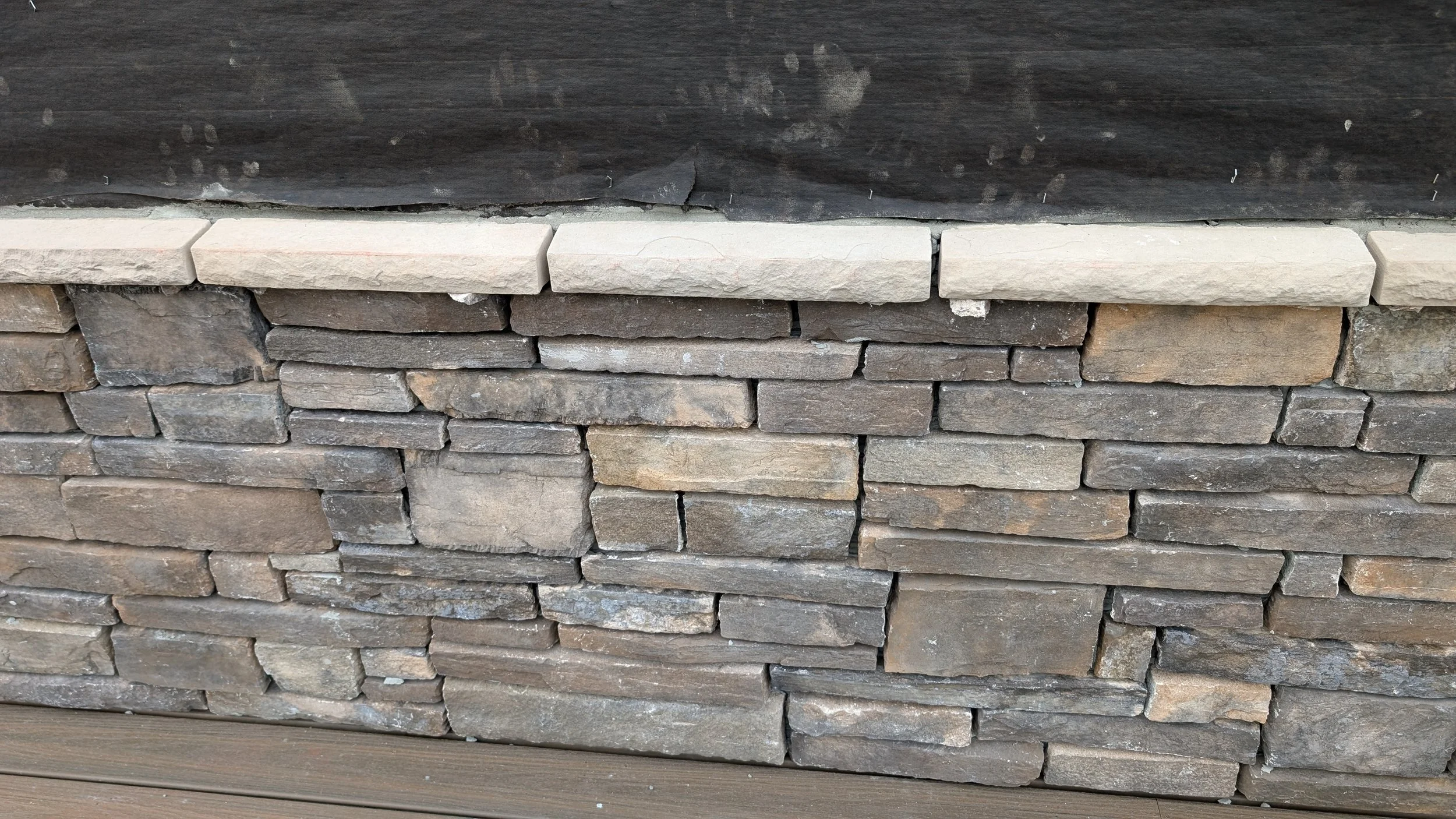 Close-up of a stone wall with layered, rectangular stones of various sizes and colors, topped with flat beige stone. The wall is below a dark surface or roof edge.