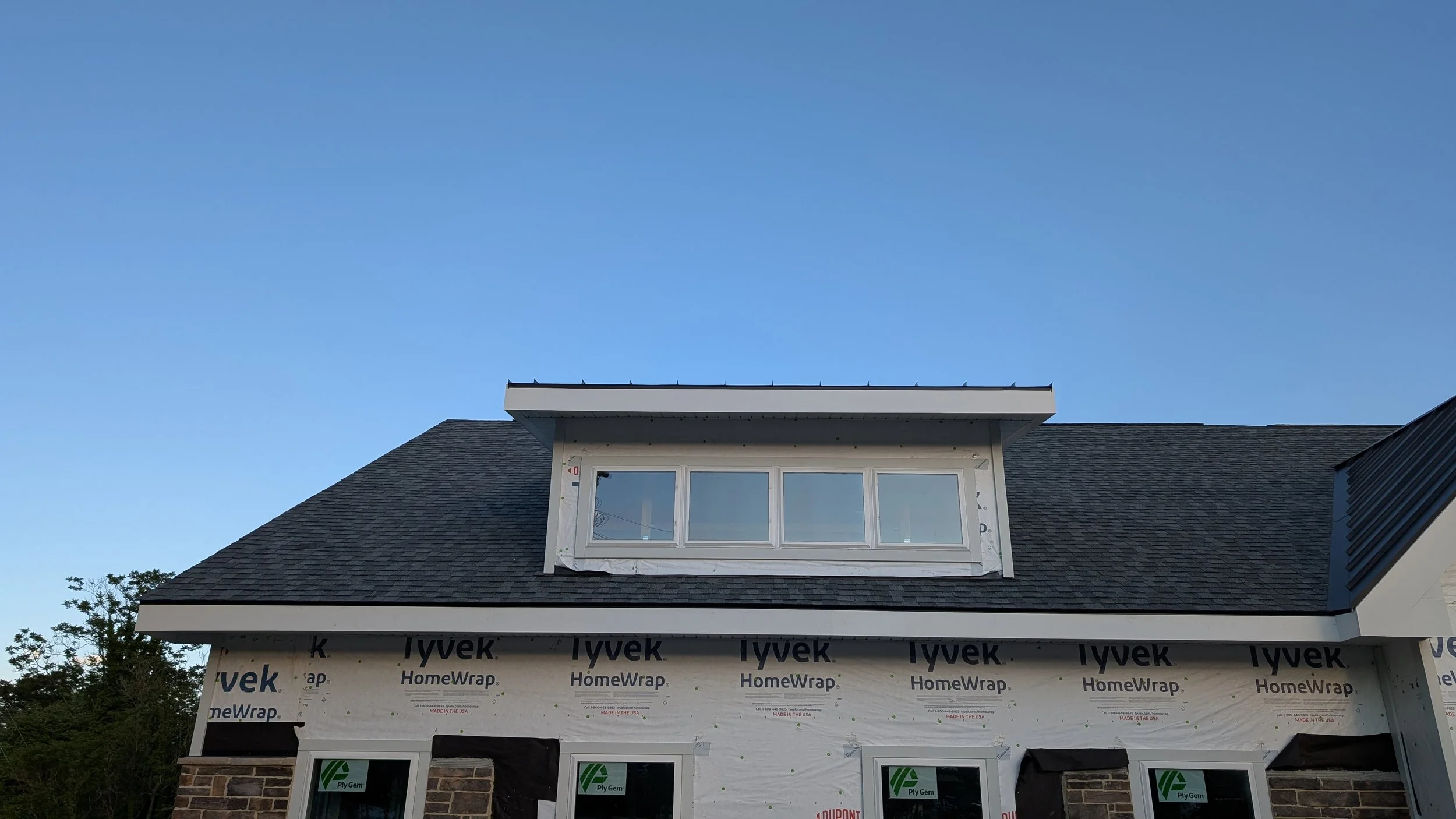 A house under construction with an unfinished upper floor. The roof is dark shingles, and there's a large window installed in the dormer. The lower part of the house has brickwork and window frames covered with protective film. The sky is clear and b