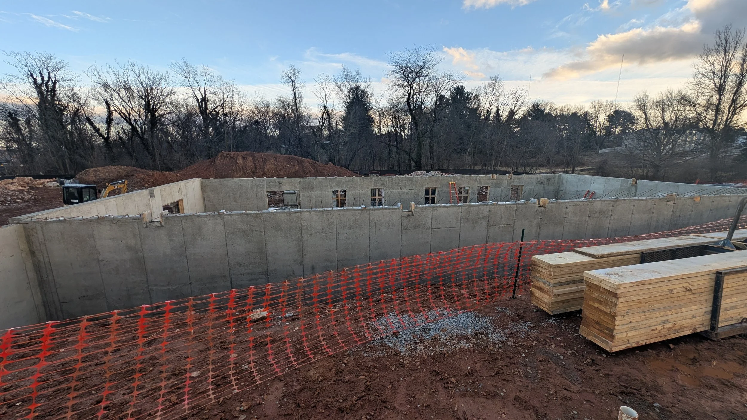 Construction site with concrete walls and lumber stacks, orange safety fencing, and trees in the background under a partly cloudy sky.