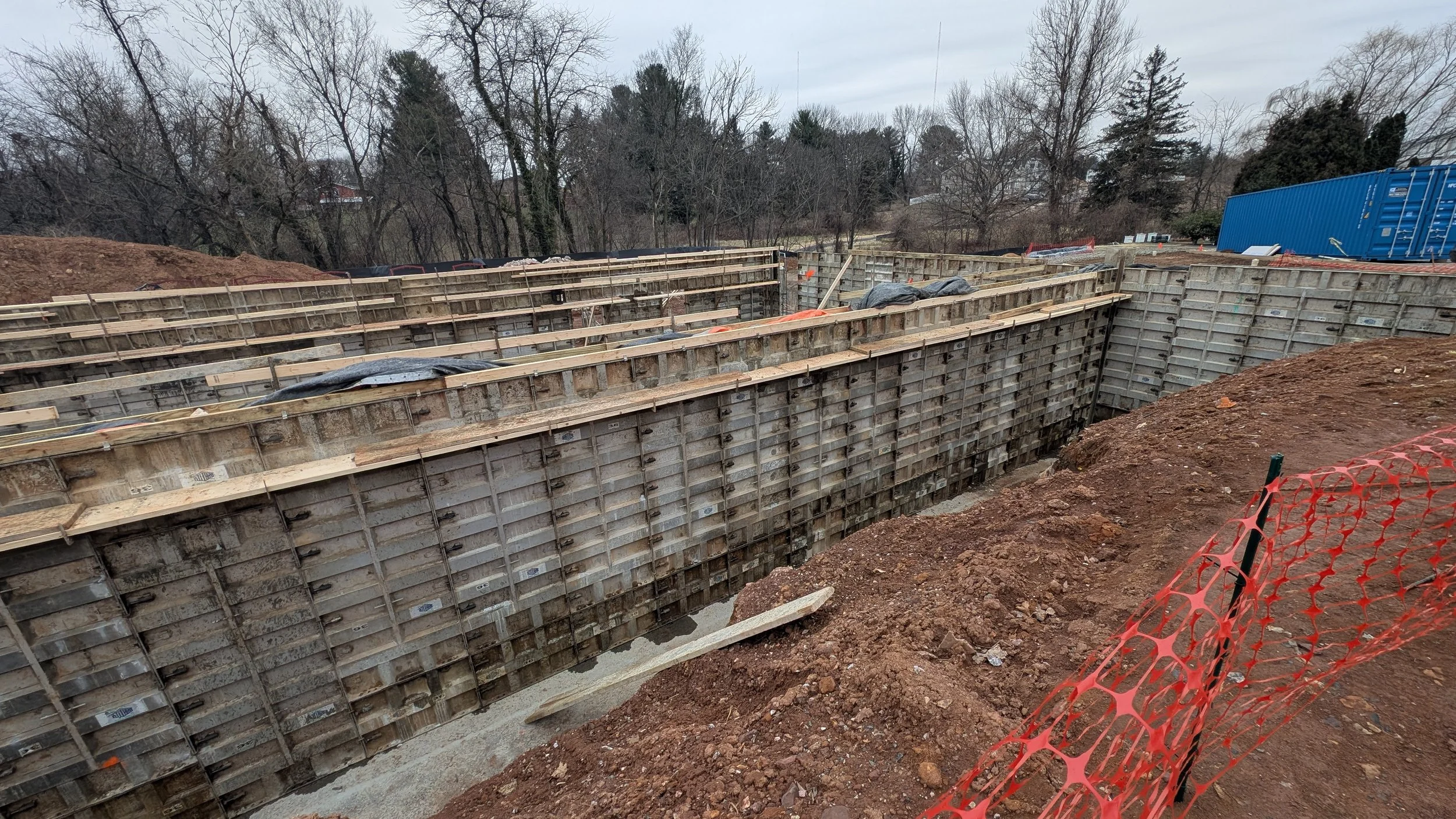 Construction site with concrete formwork and wooden supports for a foundation, red dirt, and orange safety fencing.