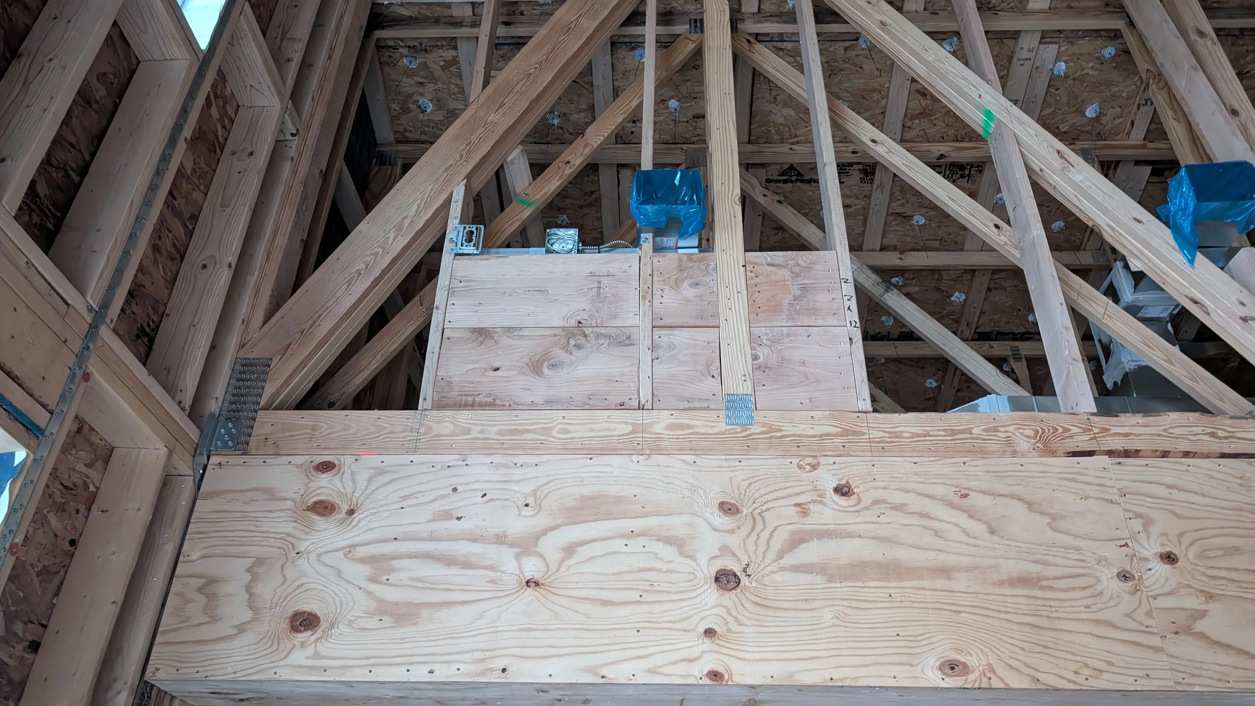 Interior view of an unfinished attic or upper floor under construction, with exposed wooden framing, wall studs, and partially installed plywood flooring. Some construction materials and tools are visible.