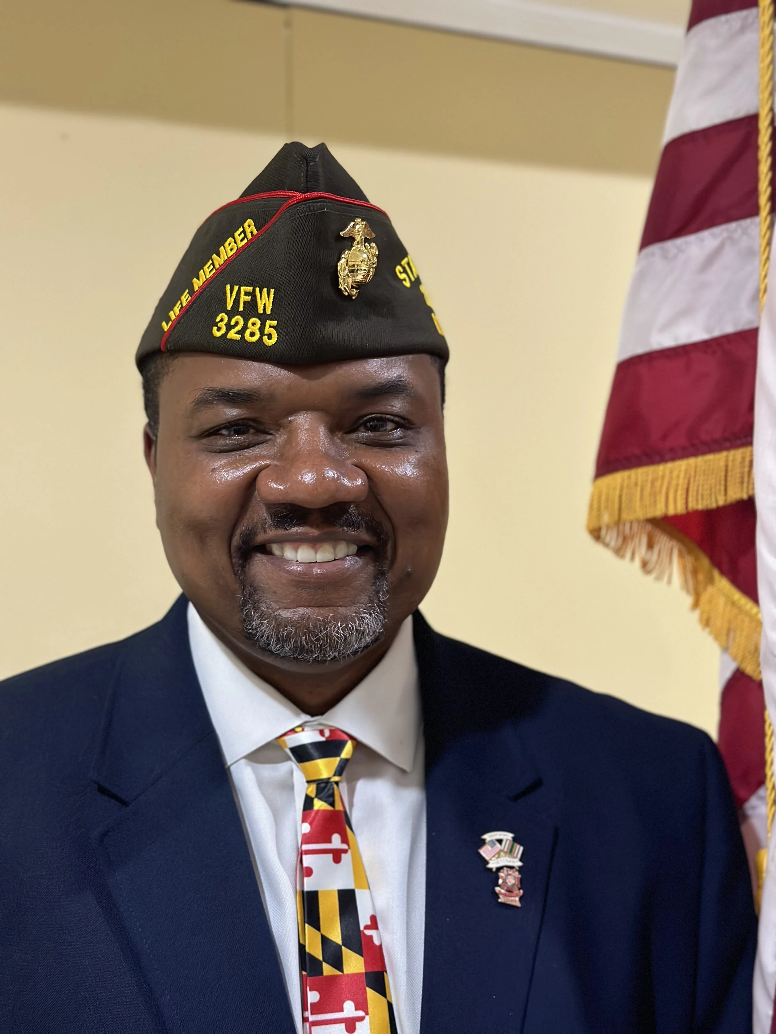 A man in a navy suit and white shirt, wearing a black military-style hat with yellow embroidery reading 'Life Member VFW 3285' and a Marine Corps emblem. He has a patriotic tie with Maryland flag colors and a pin resembling a flag on his lapel. An American flag is partially visible on the right side of the image.