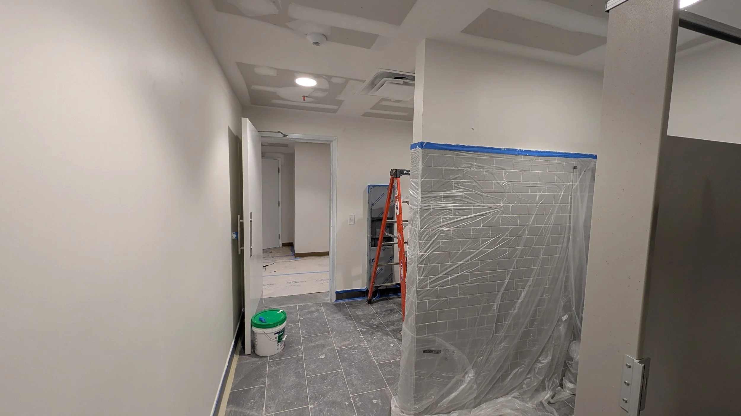 Interior view of a building under renovation with a ladder, plastic-covered wall, painted tiles, a bucket, and a door.