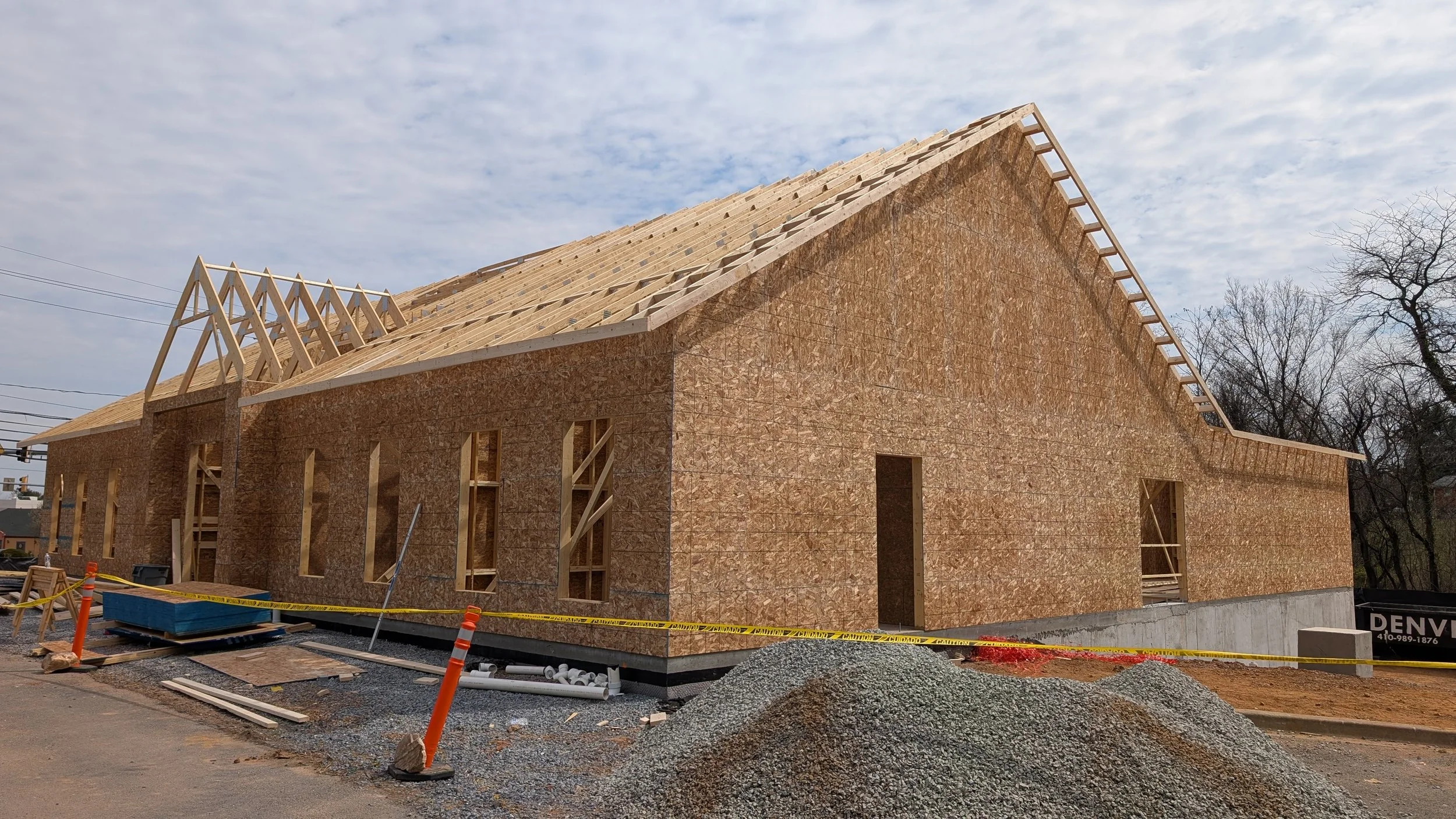 A house under construction with wooden framing and sheathing, surrounded by construction materials, orange safety cones, and yellow caution tape, with trees and a cloudy sky in the background.
