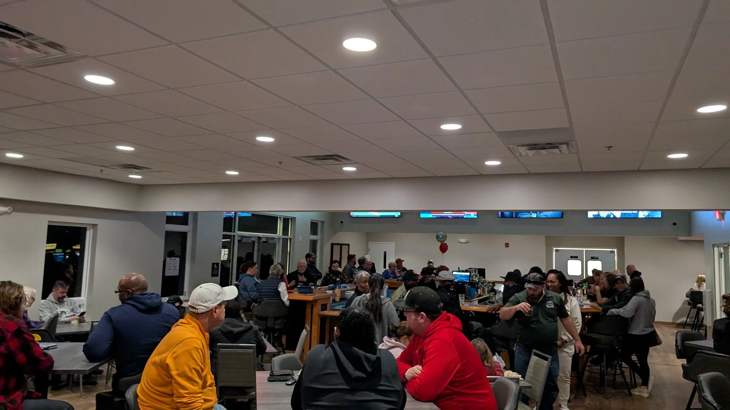 People gathering in a well-lit indoor space, sitting at tables and standing near the bar, with digital screens above and doors in the background.