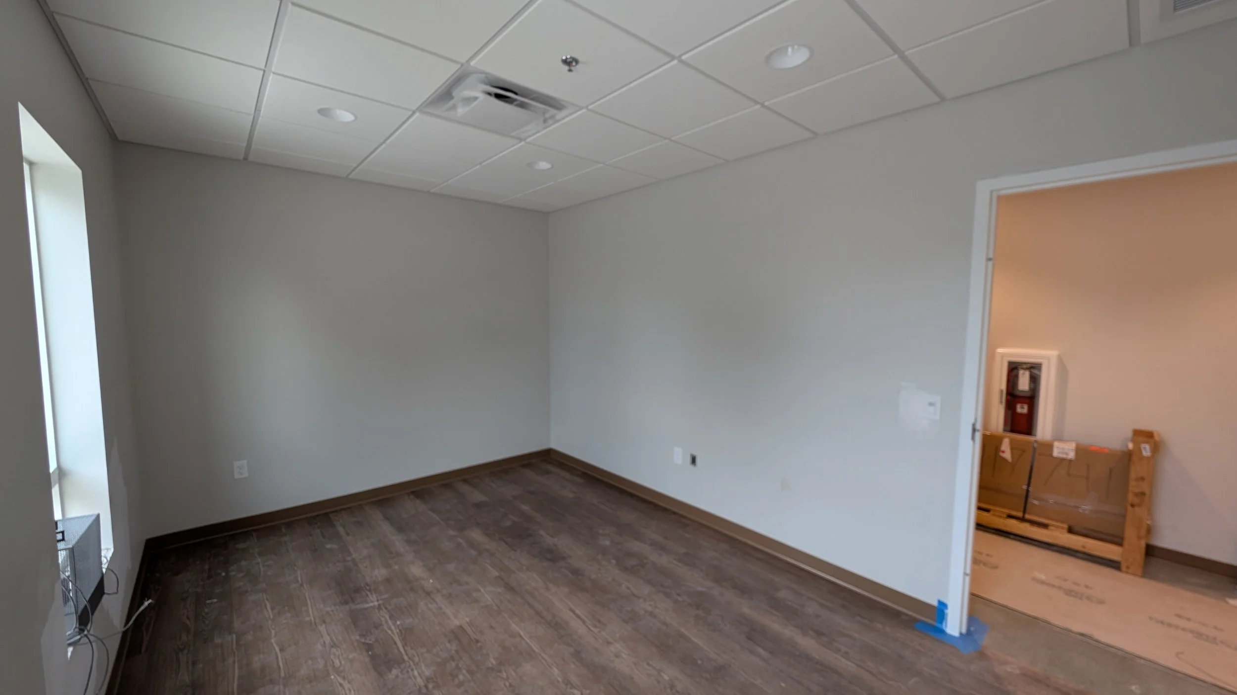An empty room with light-colored walls, a brown hardwood floor, a white ceiling with recessed lighting, and a small window on the left. There is a partially open doorway on the right leading to another room with construction materials.